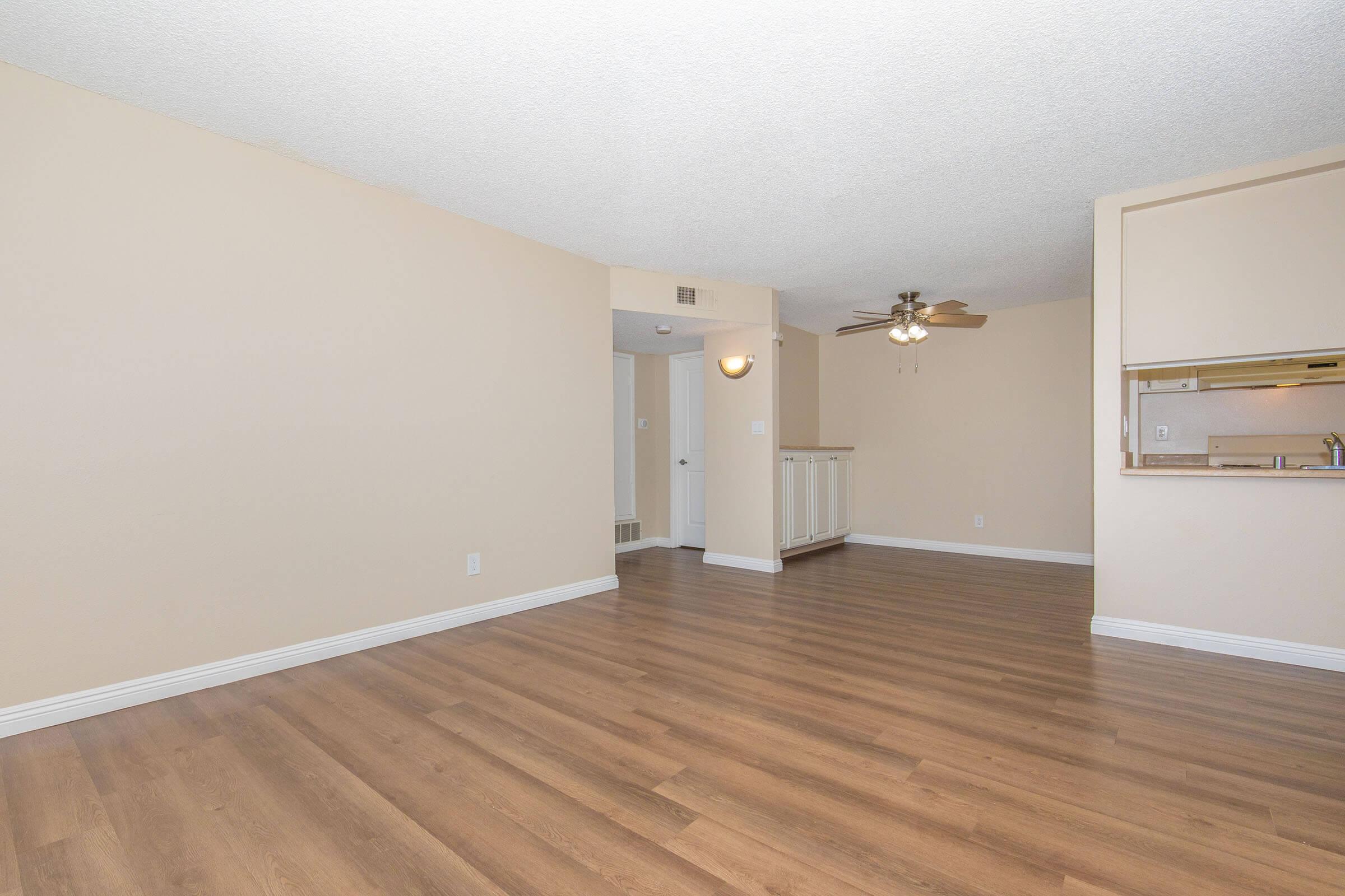 A spacious, well-lit living area with light beige walls and hardwood flooring. A ceiling fan is visible in the corner, and there is an open kitchen area in the background with a pass-through bar. The layout appears clean and inviting, perfect for a cozy living space.