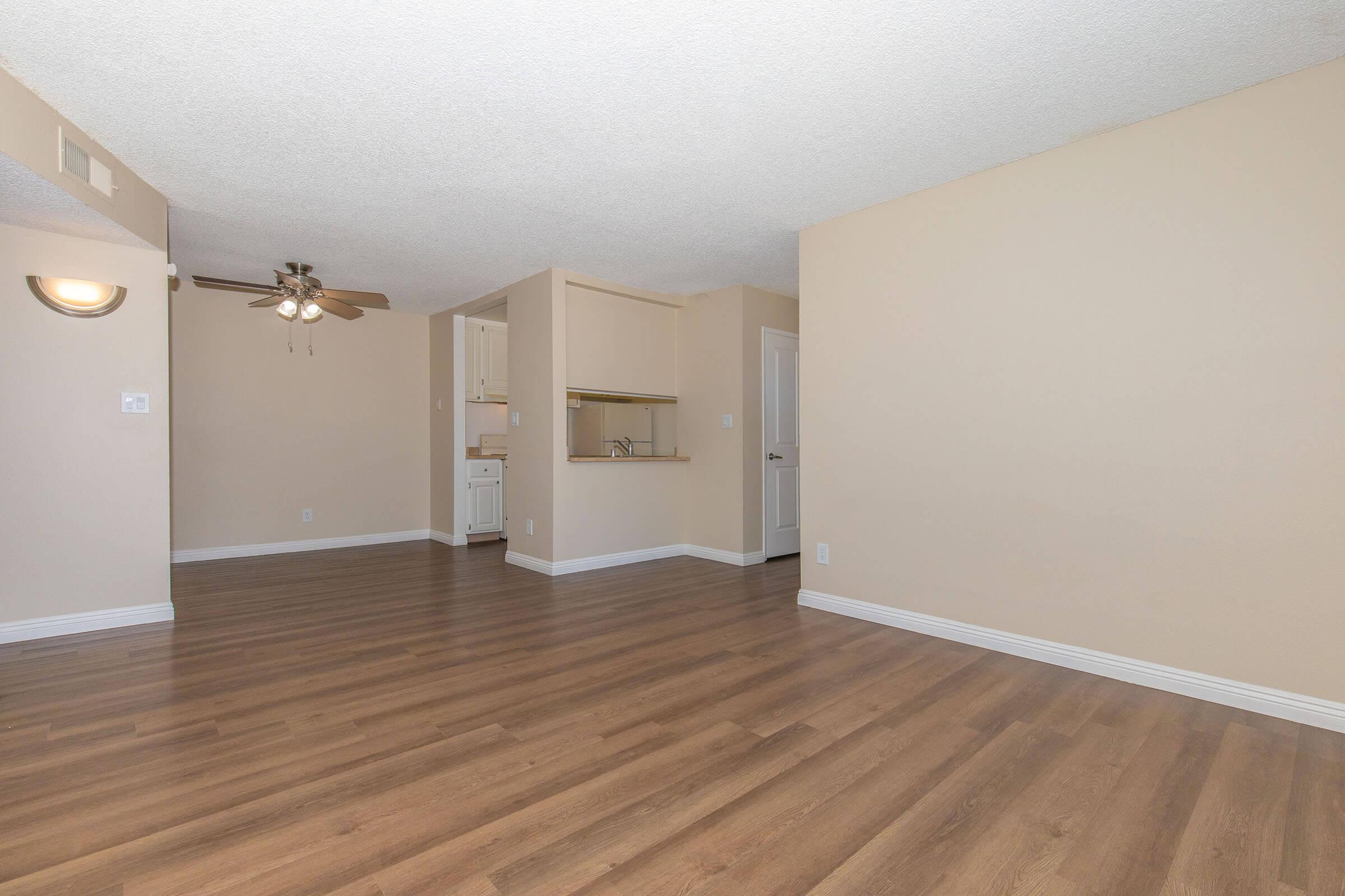 A spacious, empty living area with light-colored walls and laminate flooring. The room features a ceiling fan, with a partial view of a kitchen area in the background, which includes a bar and white cabinetry. Natural light fills the space, creating a bright and inviting atmosphere.