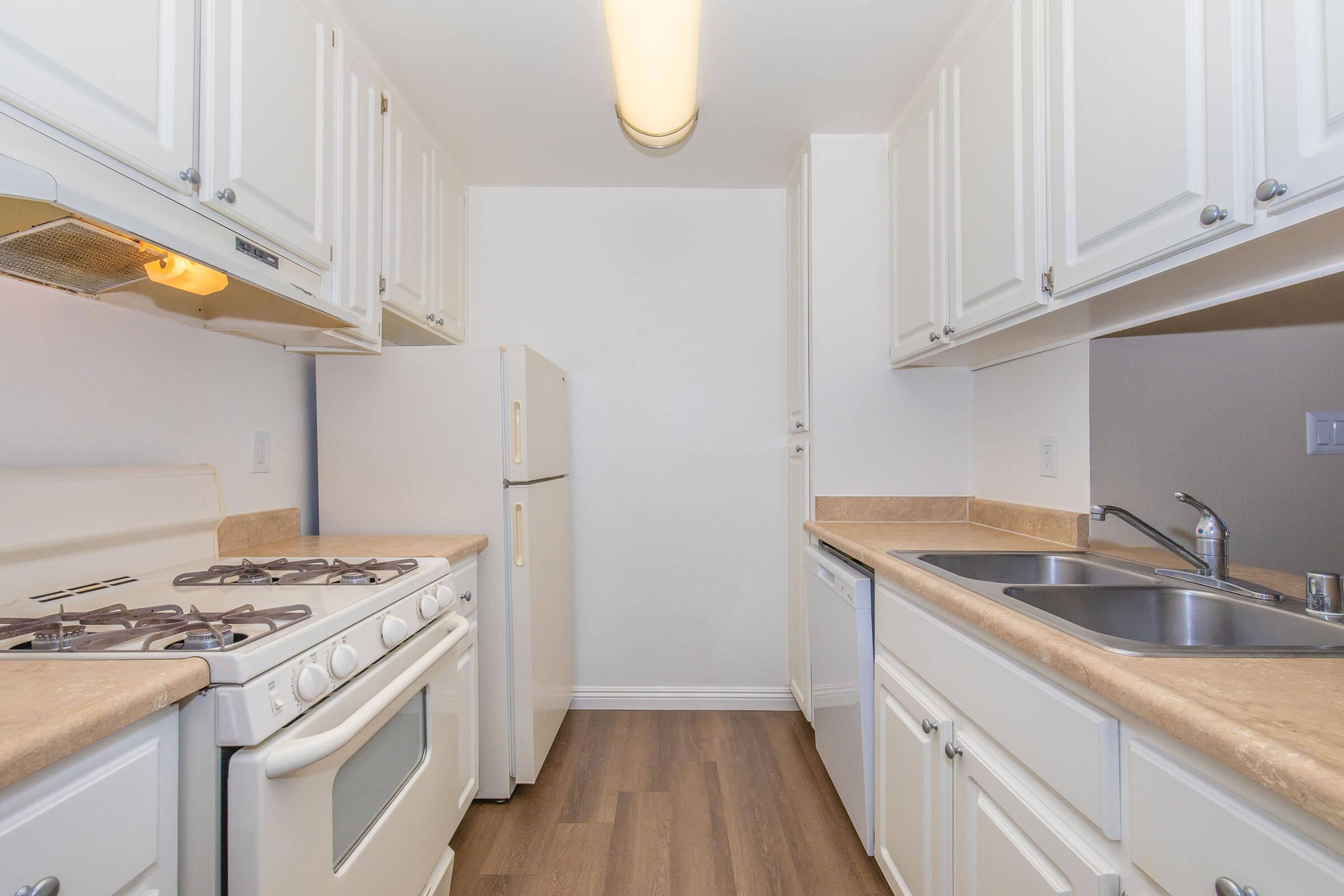 A modern kitchen featuring white cabinetry, a gas stove, refrigerator, and double sink. The countertops are beige, and the flooring is a wood laminate. The space is well-lit with a ceiling light, creating a clean and inviting atmosphere.