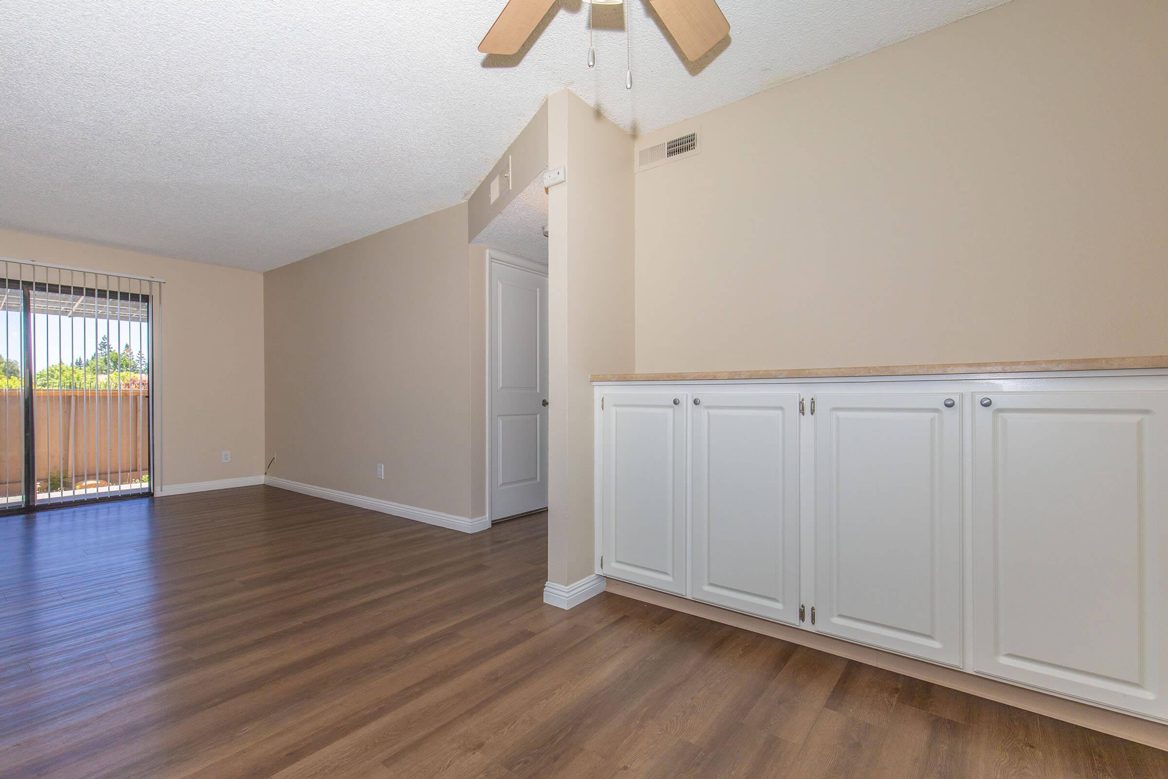 An interior view of a room with light-colored walls and wooden flooring. There is a ceiling fan visible, and a white cabinet line the wall. A door leads to an outdoor area with visible greenery, and there are large windows allowing natural light into the space.