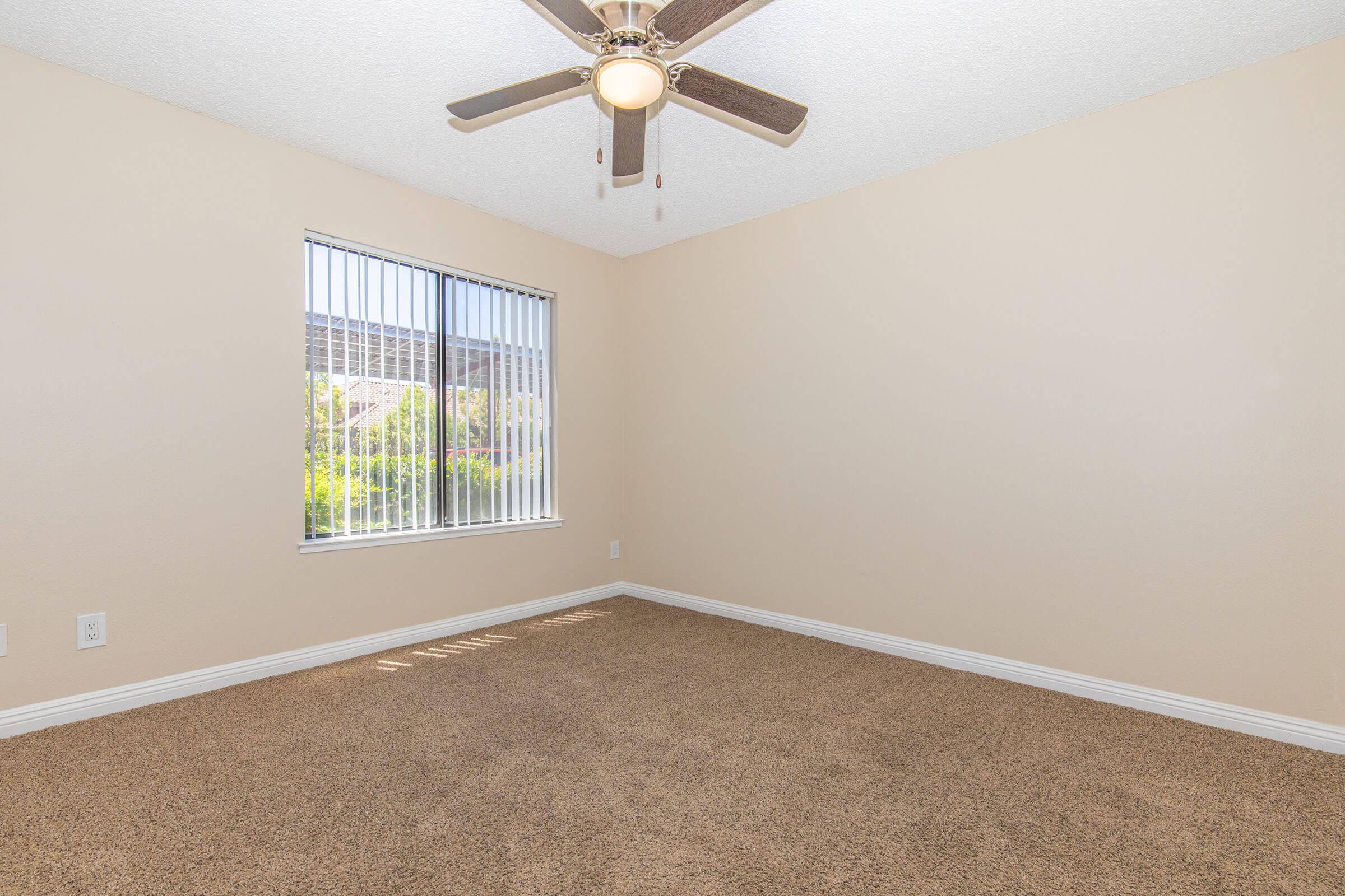 A spacious, empty room featuring light beige walls and carpet, with a ceiling fan in the center. A window with vertical blinds allows natural light to enter, casting shadows on the floor. The room is clean and ready for furnishing, creating a bright and inviting atmosphere.