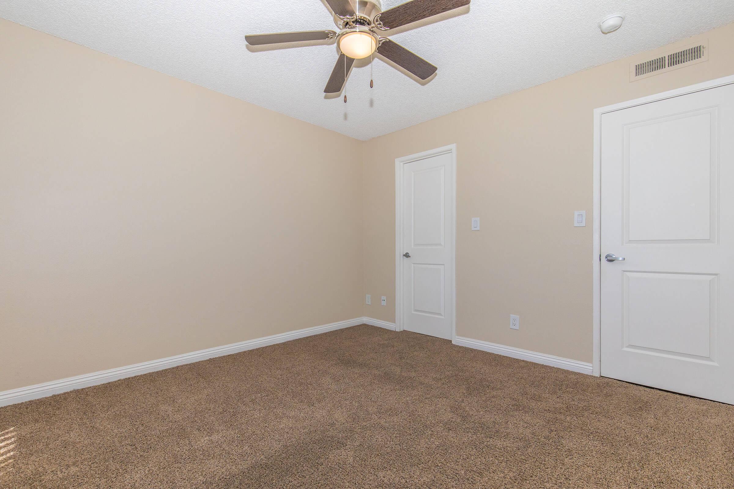 A spacious, empty bedroom featuring light beige walls, a ceiling fan with wooden blades, and plush brown carpet. Two white doors are visible: one leading to a closet and the other to the hallway. Natural light streams in, creating a bright and airy atmosphere.