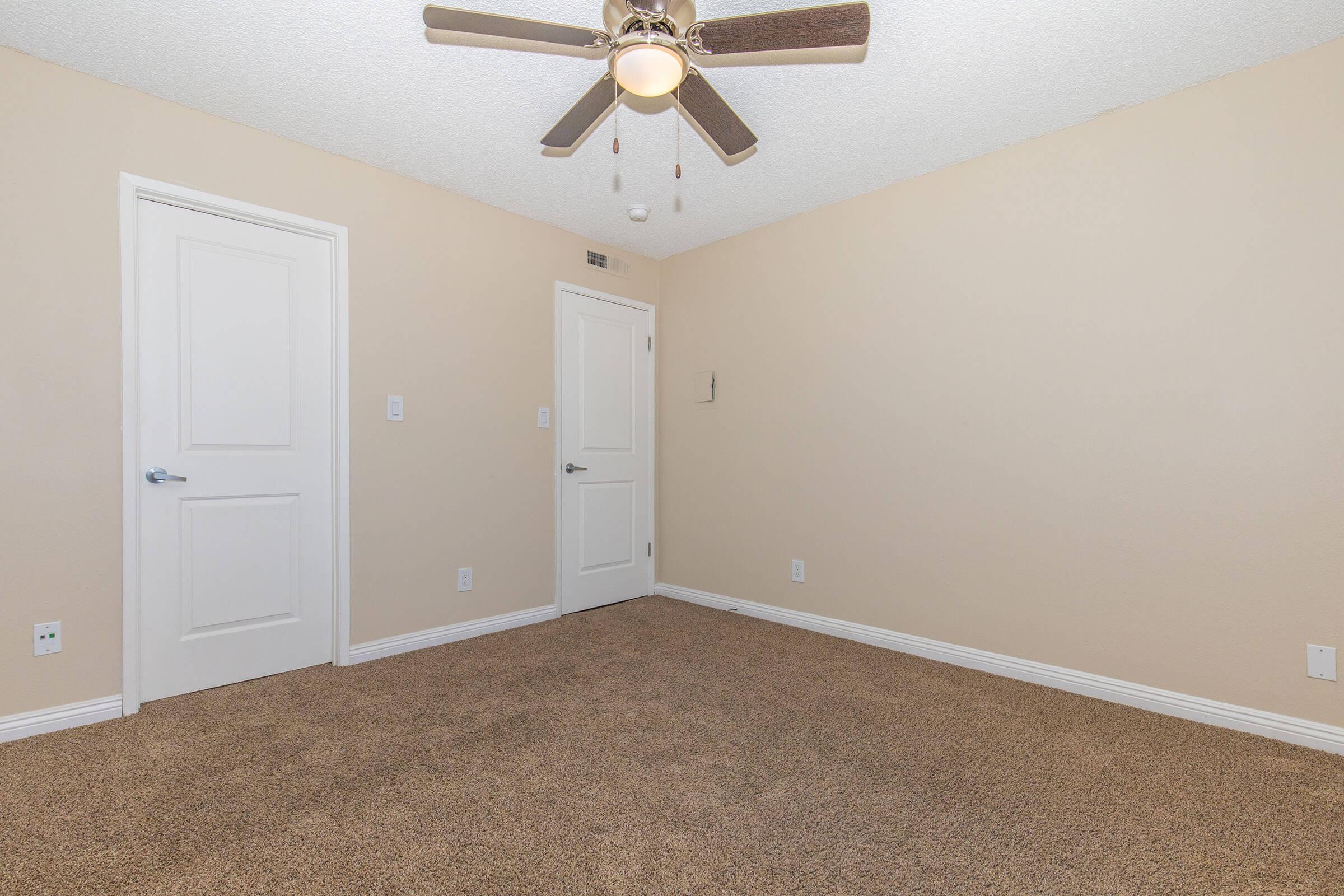 A spacious, empty room featuring beige walls and carpeted flooring. There is a ceiling fan with wooden blades, and two white doors leading to different areas. The room is bright with natural light, creating a welcoming and neutral space, ready for furniture and personal touches.