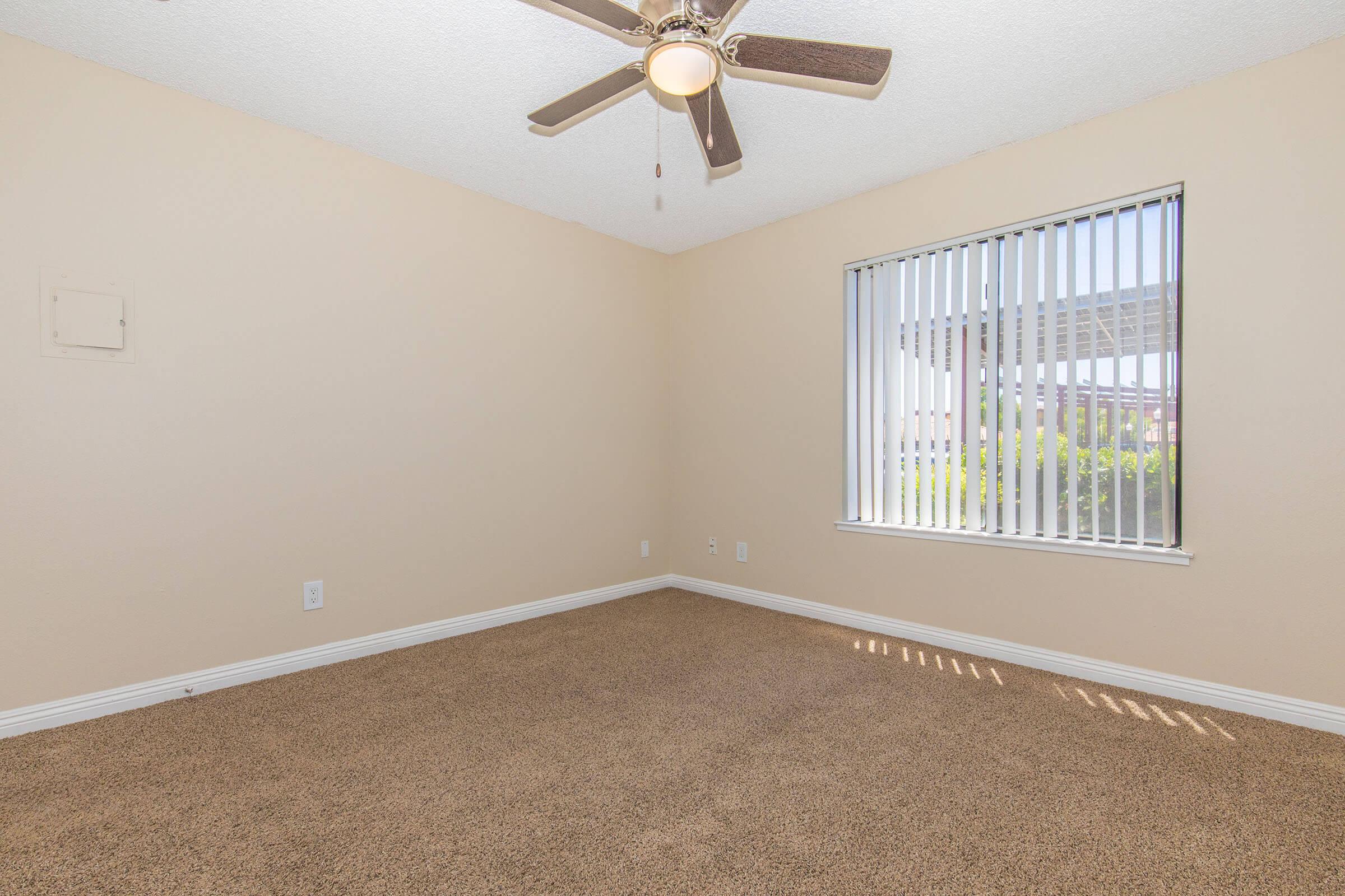 A spacious, empty room with light brown carpet and beige walls. It features a ceiling fan with wooden blades and a window covered with vertical blinds, allowing sunlight to enter. The room has a simple design, offering a blank canvas for potential furnishings and decoration.