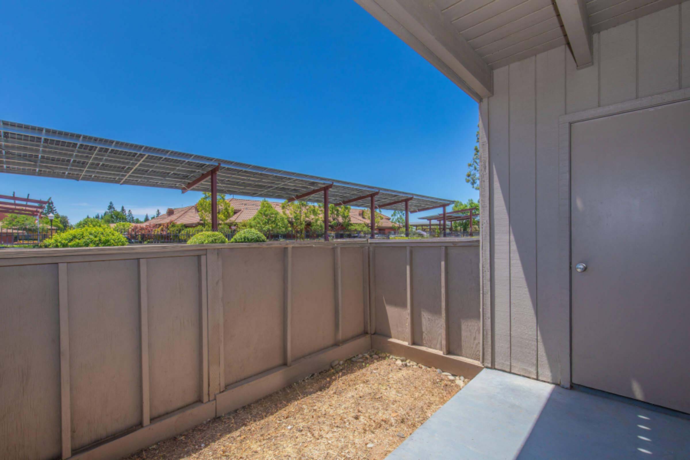 A corner view of a small outdoor space with gray walls and a sliding door. In the background, there are solar panels and several trees. The sky is clear and blue, indicating a sunny day. The ground is bare with some dirt, suggesting the area is not fully landscaped.