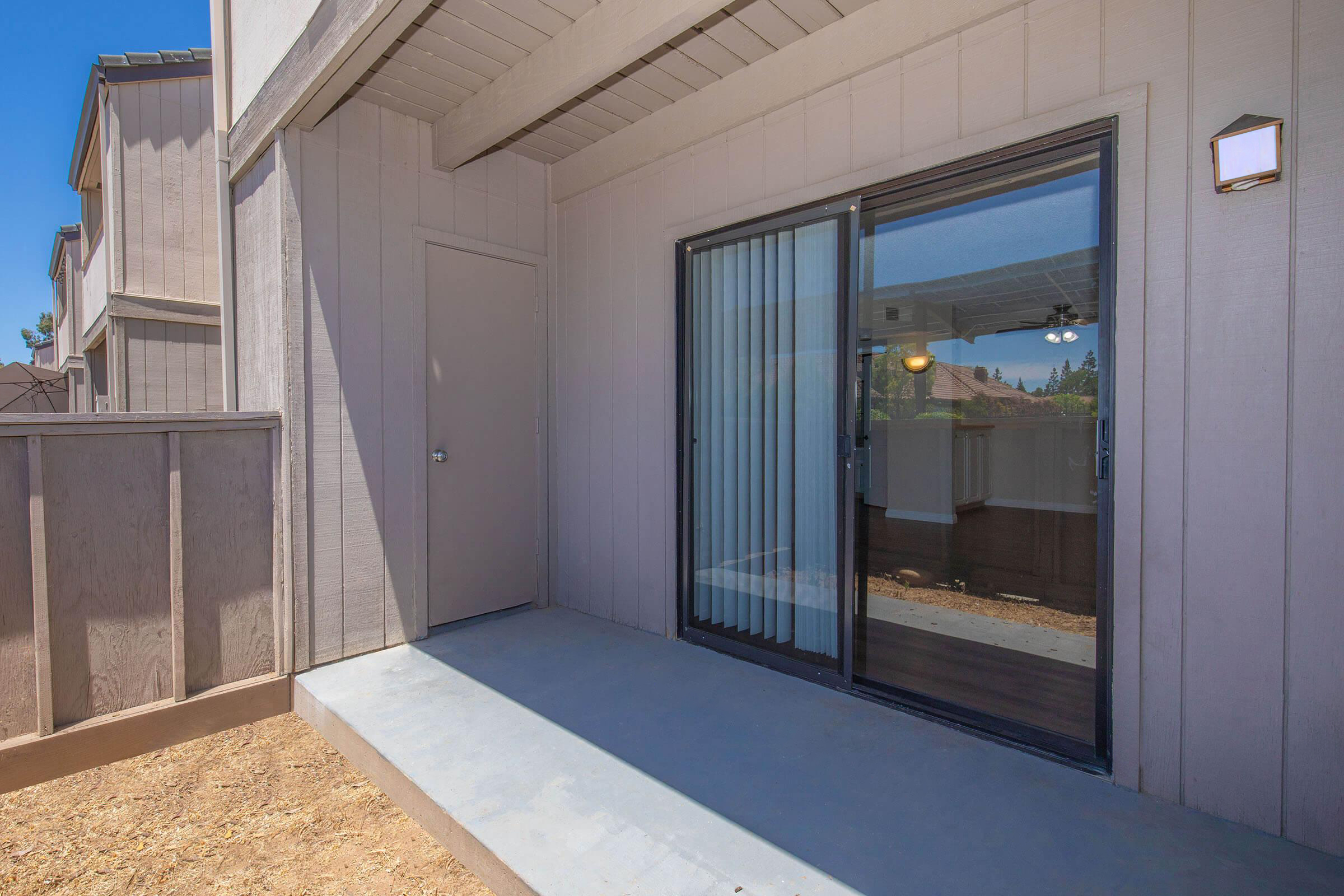 A spacious outdoor patio area with a sliding glass door leading inside, framed by light-colored walls. The concrete floor is clean and dry, and there is a small light fixture next to the door. The view is partially obscured by a fence, suggesting a private outdoor space.