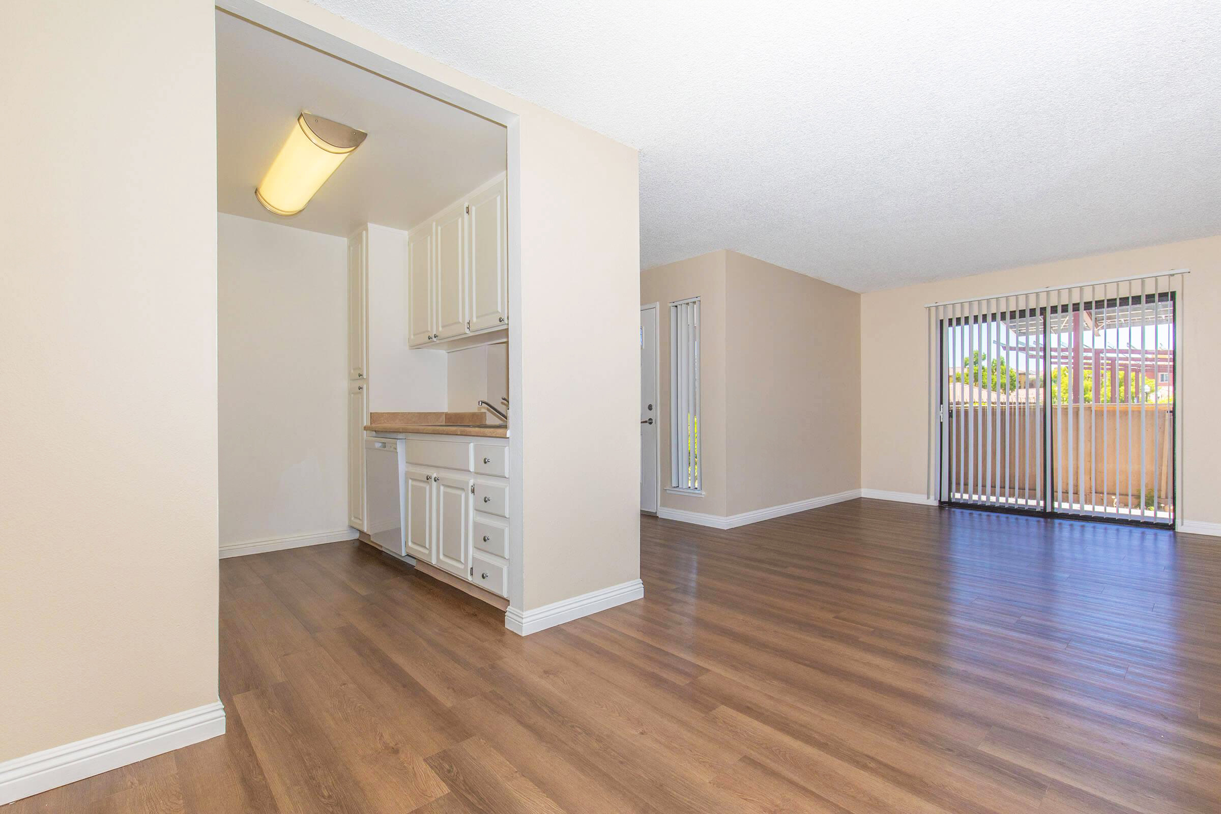 An empty room with light-colored walls and wooden flooring. On the left, a small kitchenette with cabinets and a countertop. To the right, a spacious living area with large windows covered by vertical blinds, allowing natural light to enter.
