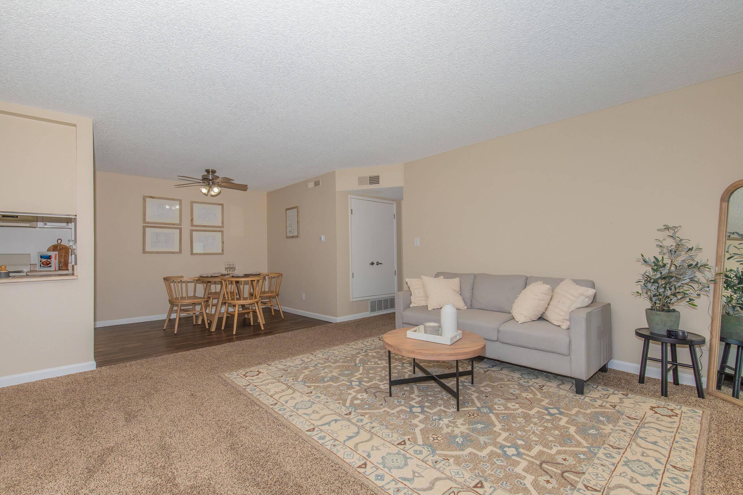 A cozy living room featuring a light gray sofa with cushions, a round coffee table, and a decorative plant. Nearby, a dining area with a wooden table and chairs is visible. The space has beige walls, carpeted flooring, and subtle lighting, creating a warm and inviting atmosphere.