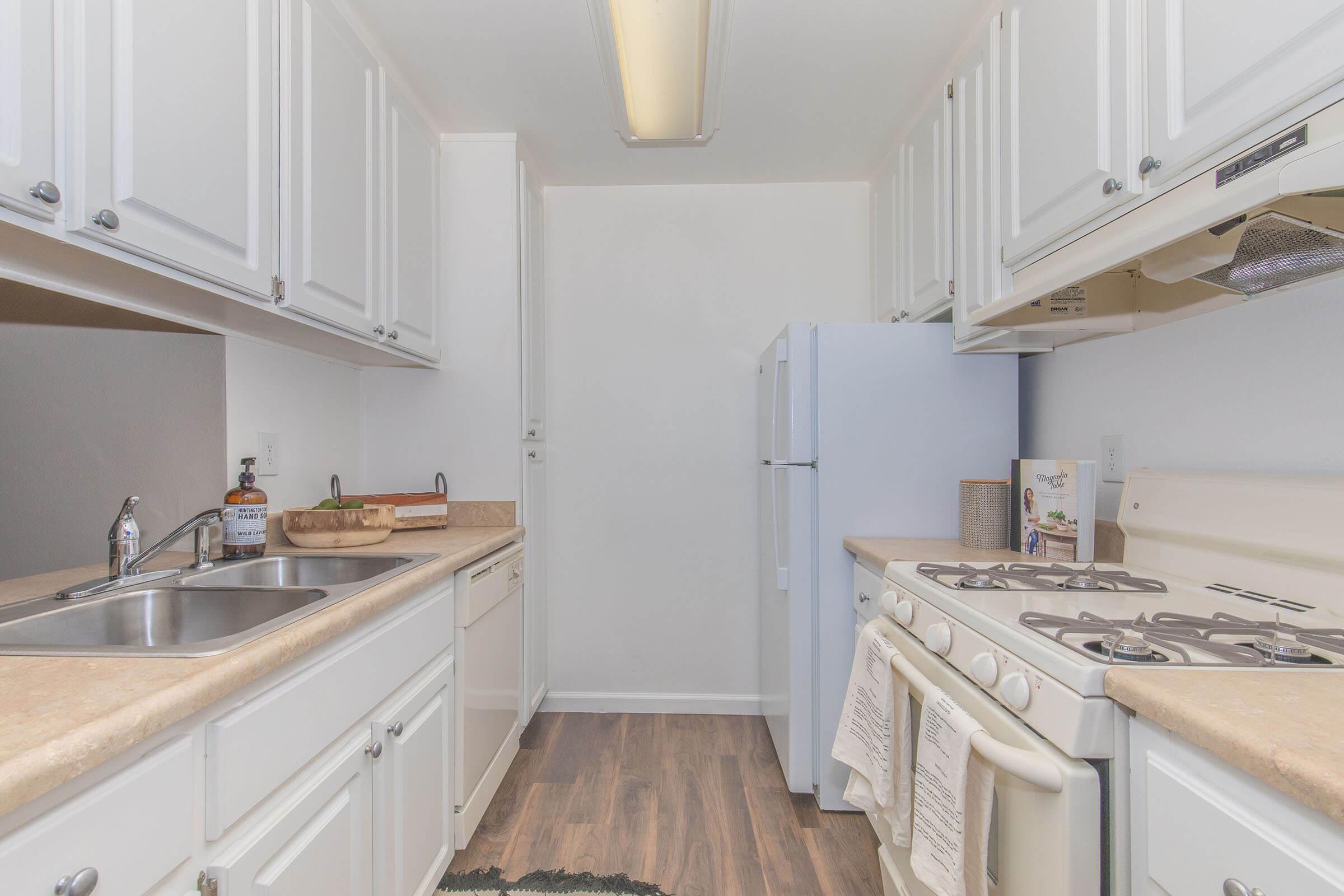 A bright and modern kitchen featuring white cabinets, a double sink, a beige countertop, a white refrigerator, and a gas stove. The space is well-organized with minimal decor, showcasing a clean and inviting atmosphere.
