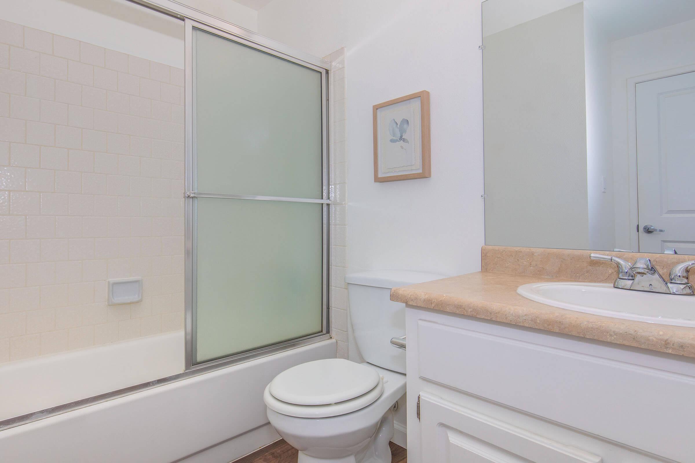 A clean, modern bathroom featuring a bathtub with a transparent sliding glass door, a white toilet, and a countertop sink. A framed botanical print is on the wall, and natural light enhances the bright, neutral color scheme of the space. The flooring has a wooden appearance.