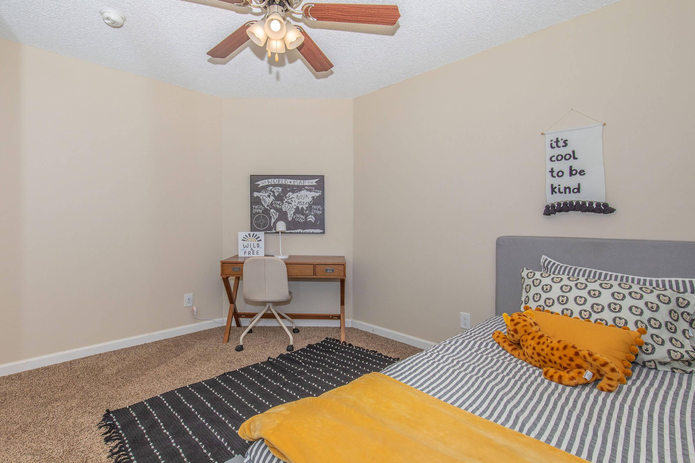 A cozy bedroom featuring a bed with patterned linens, a yellow throw blanket, and a stuffed tiger toy. There’s a small desk with a chair, a black and white art print on the wall, and a wall banner that reads "it's cool to be kind." The room has light-colored walls and a ceiling fan.