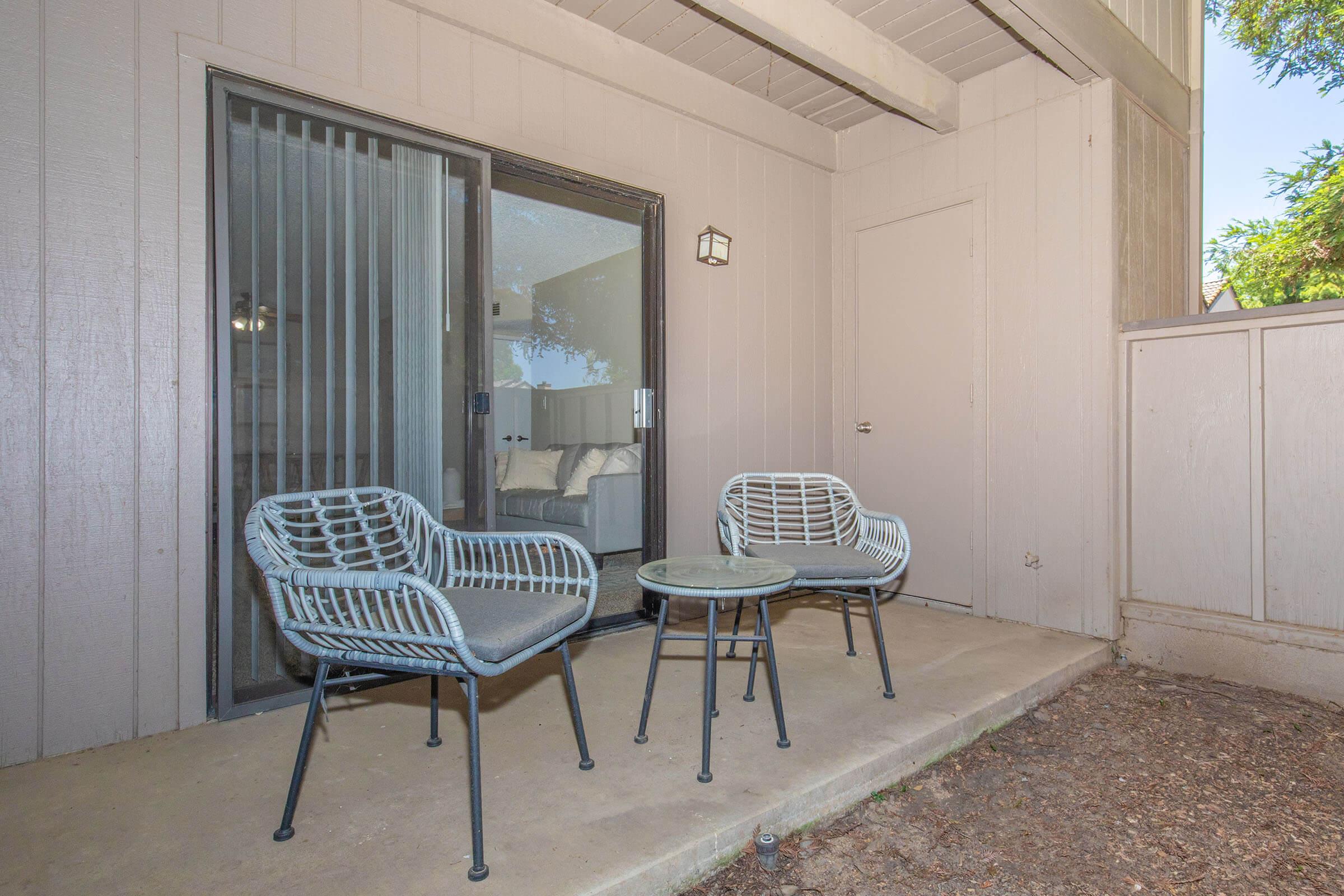 A small outdoor patio area featuring two decorative chairs and a glass-topped table, set against a backdrop of a sliding glass door leading inside. The atmosphere is calm and inviting, surrounded by a light-colored wall and greenery in the background.