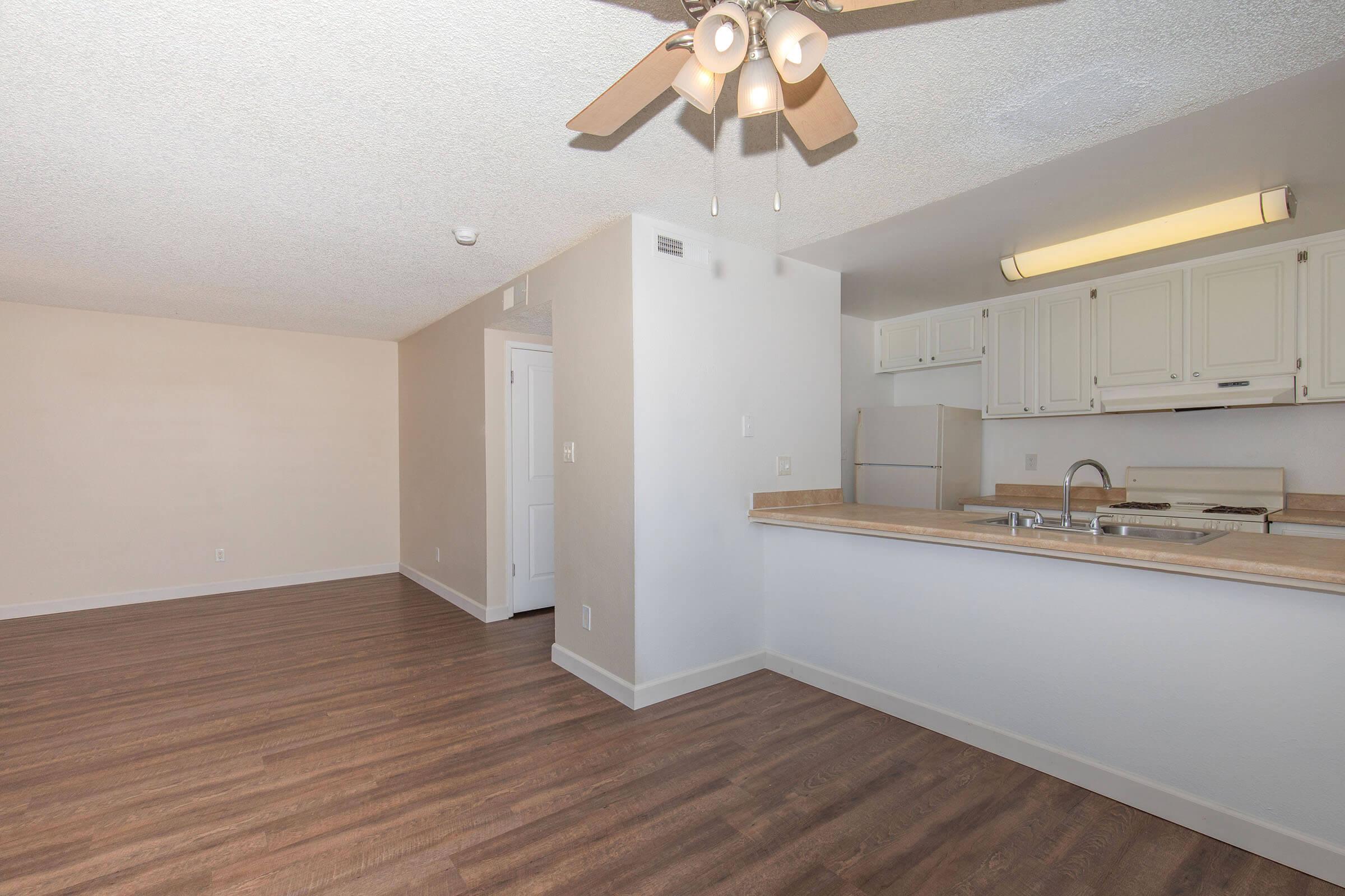 A spacious, well-lit room with light-colored walls and a wooden floor. There is a kitchen area with white cabinets and appliances, a countertop with a sink, and a ceiling fan overhead. The room has an open layout, creating a warm and inviting atmosphere.