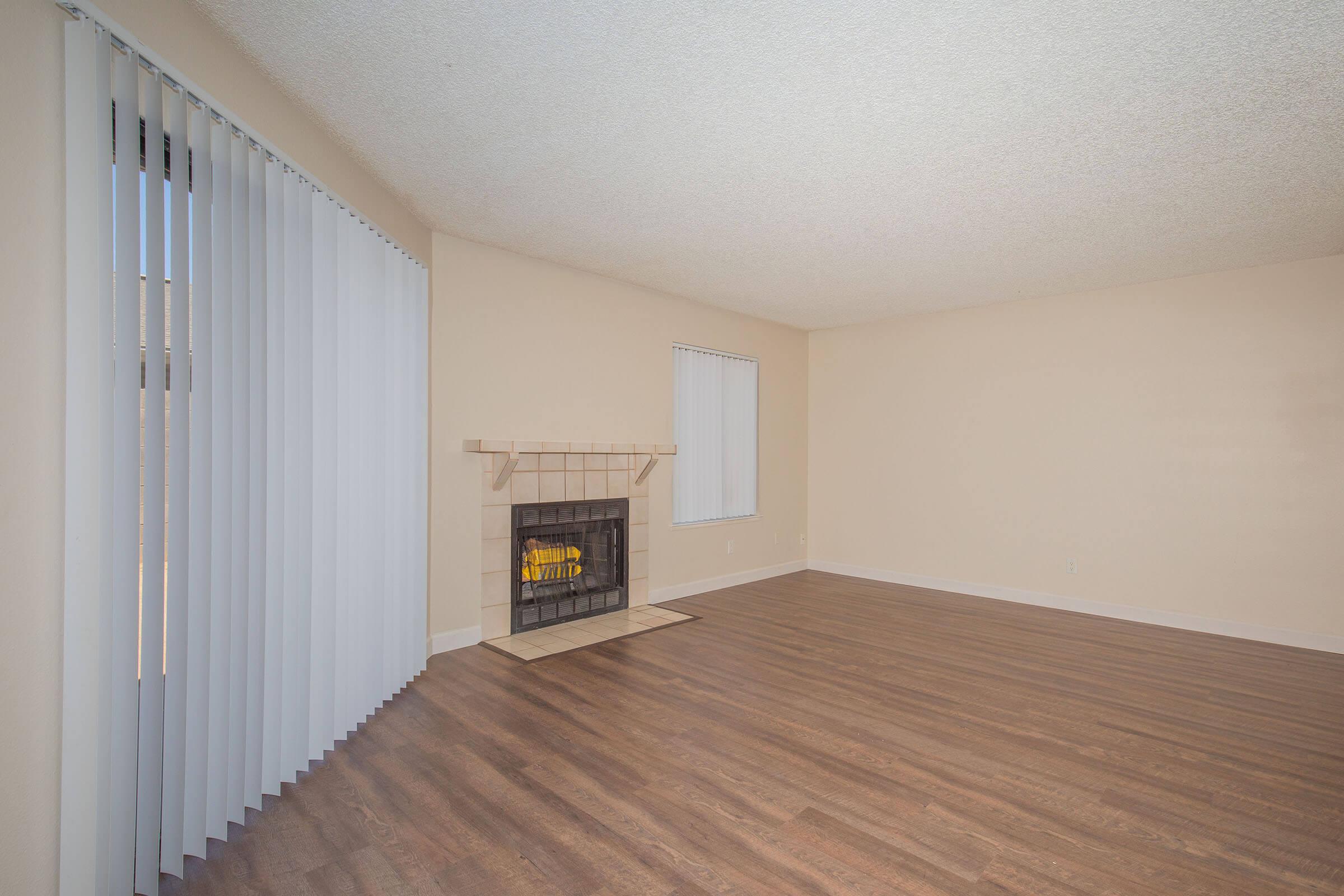 Spacious living room with light-colored walls, hardwood flooring, and a large window with vertical blinds. A brick fireplace is centrally located against one wall, and the room is empty, giving a clean and open ambiance. Natural light brightens the area, enhancing its inviting feel.