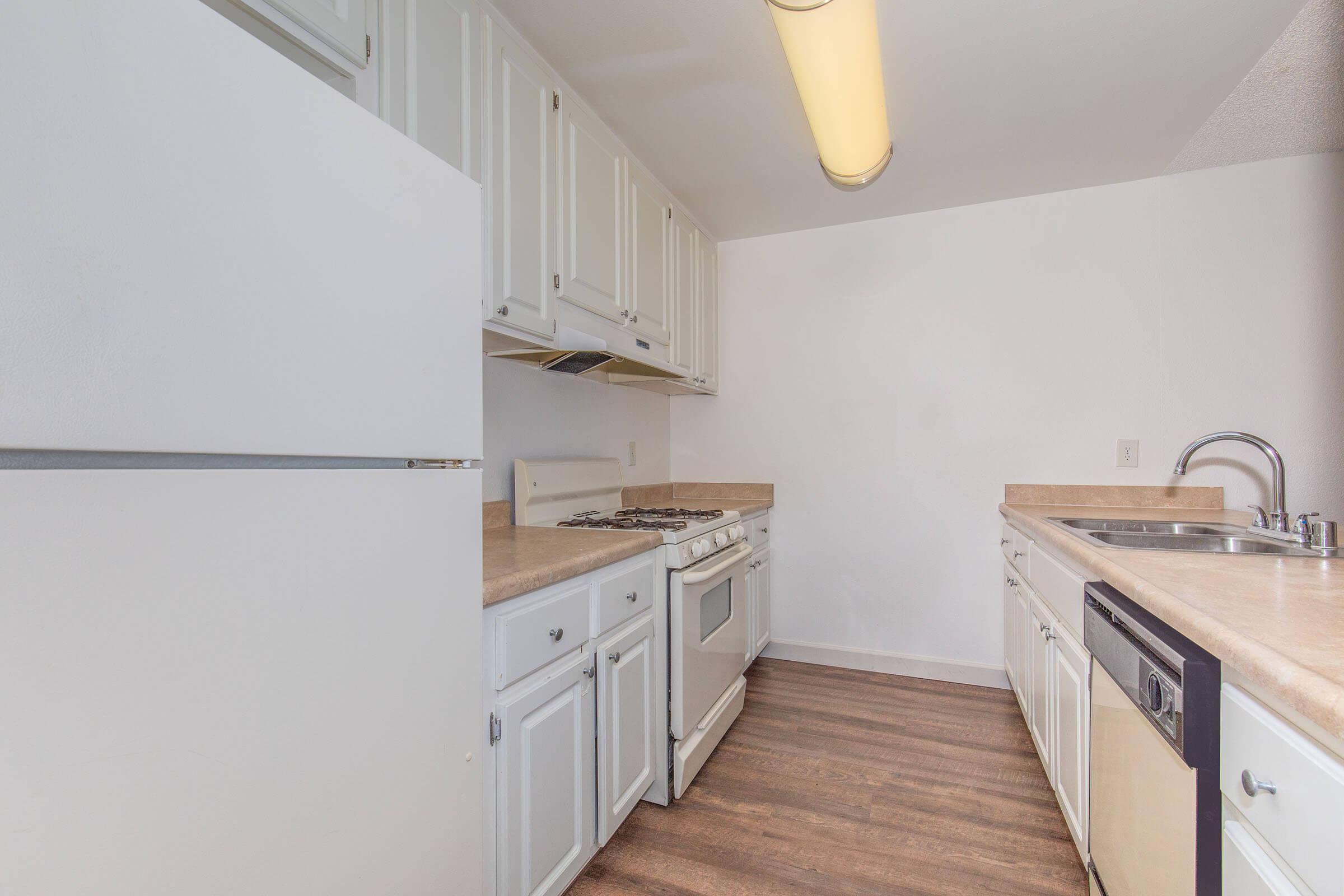 A modern kitchen featuring white cabinets, a light countertop, a gas stove, and a sink. There's a refrigerator on the left and a dishwasher next to the sink. The floor has a wood-like finish, and the overall design is functional and clean with bright lighting.
