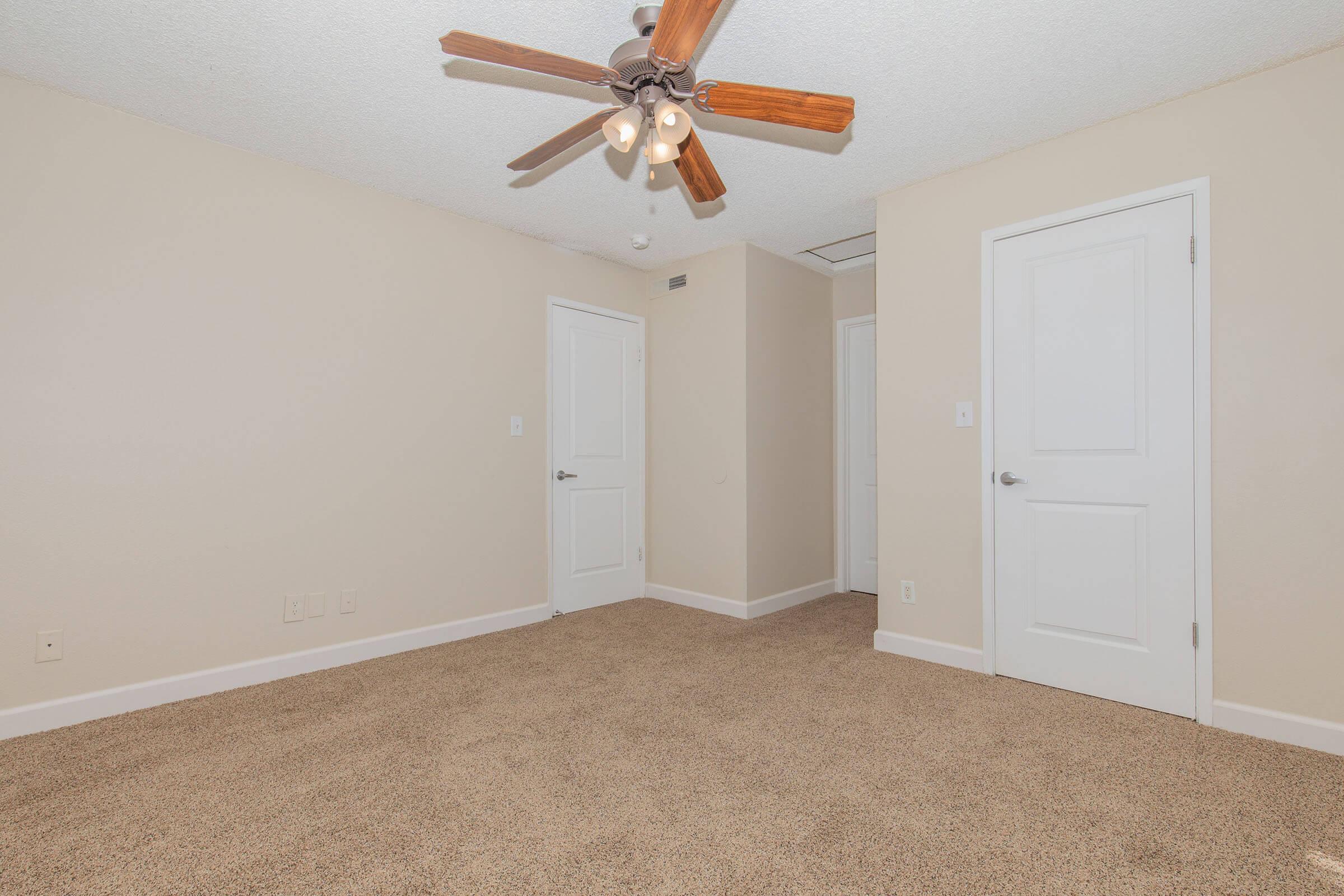 A well-lit, empty room featuring beige walls and light brown carpet. There is a ceiling fan with wooden blades, and two white doors on the left side. A small hallway can be seen leading to another door at the back. The overall ambiance is clean and spacious, suitable for various purposes.