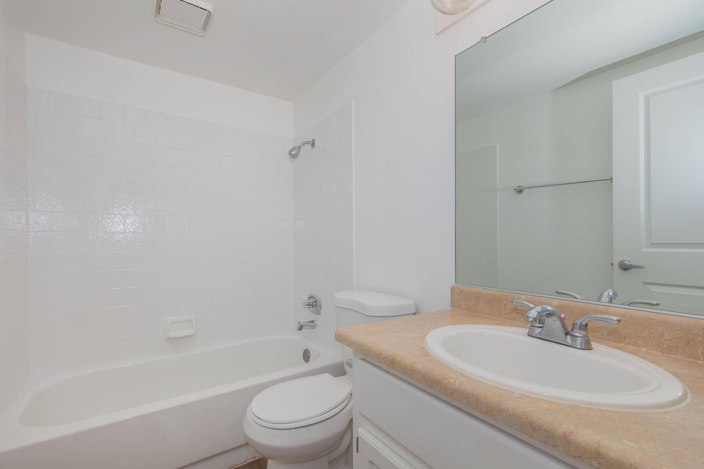 A clean and modern bathroom featuring a white tiled shower and bathtub, a beige countertop with a sink, a circular mirror above the sink, and a toilet. The walls are painted white, and there is a towel rack mounted on one side. The overall design is bright and minimalist.