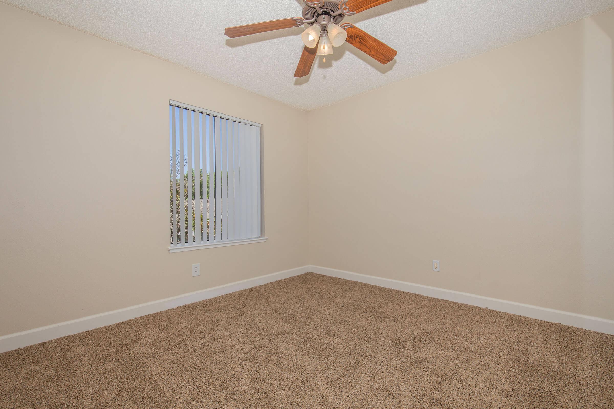 A clean, empty room featuring beige walls, carpeted flooring, and a ceiling fan with wooden blades. A window with vertical blinds allows natural light to enter the space. The overall appearance is bright and inviting, ideal for personal customization.