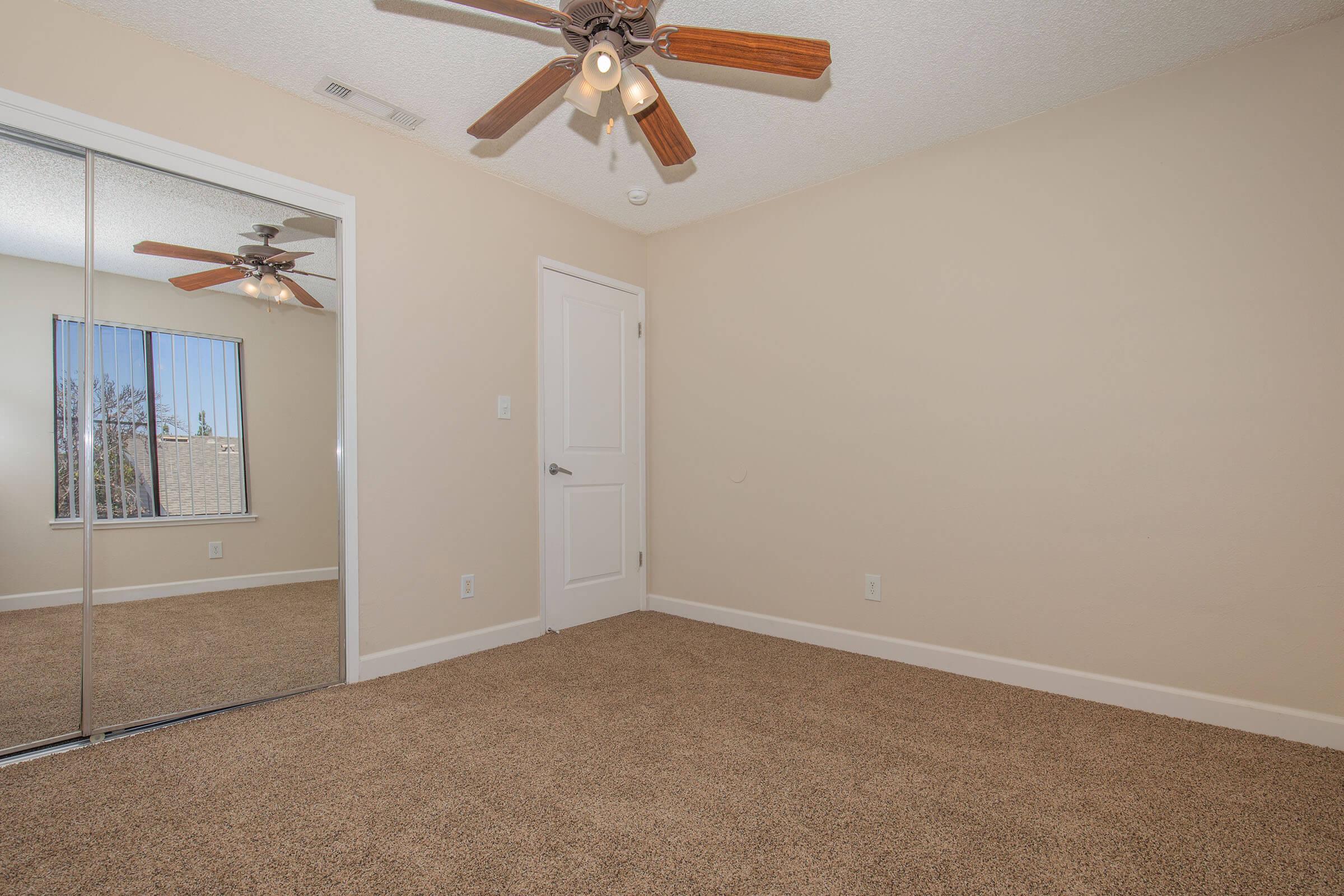 Empty room featuring a light-colored wall and beige carpet. A ceiling fan with wooden blades is installed. There is a large mirror wardrobe on one side reflecting natural light from a window. The space is clean and tidy, suitable for various purposes such as a bedroom or office.