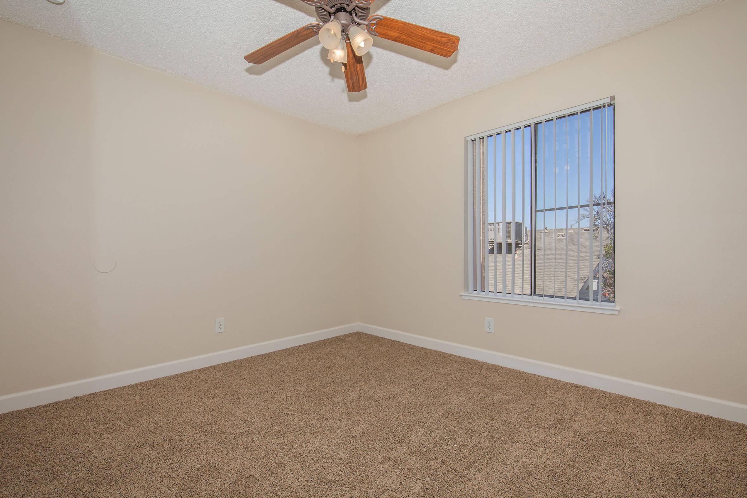 A spacious, empty room with light beige walls and a ceiling fan with wooden blades. The floor is covered in tan carpet, and a window with vertical blinds allows natural light to fill the space, providing a view of the outdoors. The overall feel is clean and inviting, perfect for personalization.