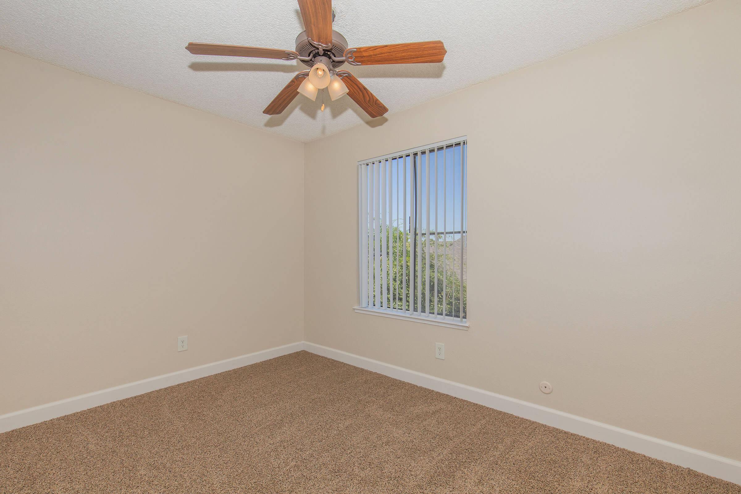 A spacious, empty room with light beige walls and a tan carpeted floor. The ceiling features a wooden ceiling fan with light fixtures, and there is a window with vertical blinds allowing natural light to enter. The room has a clean and fresh appearance, ideal for personalization or decoration.