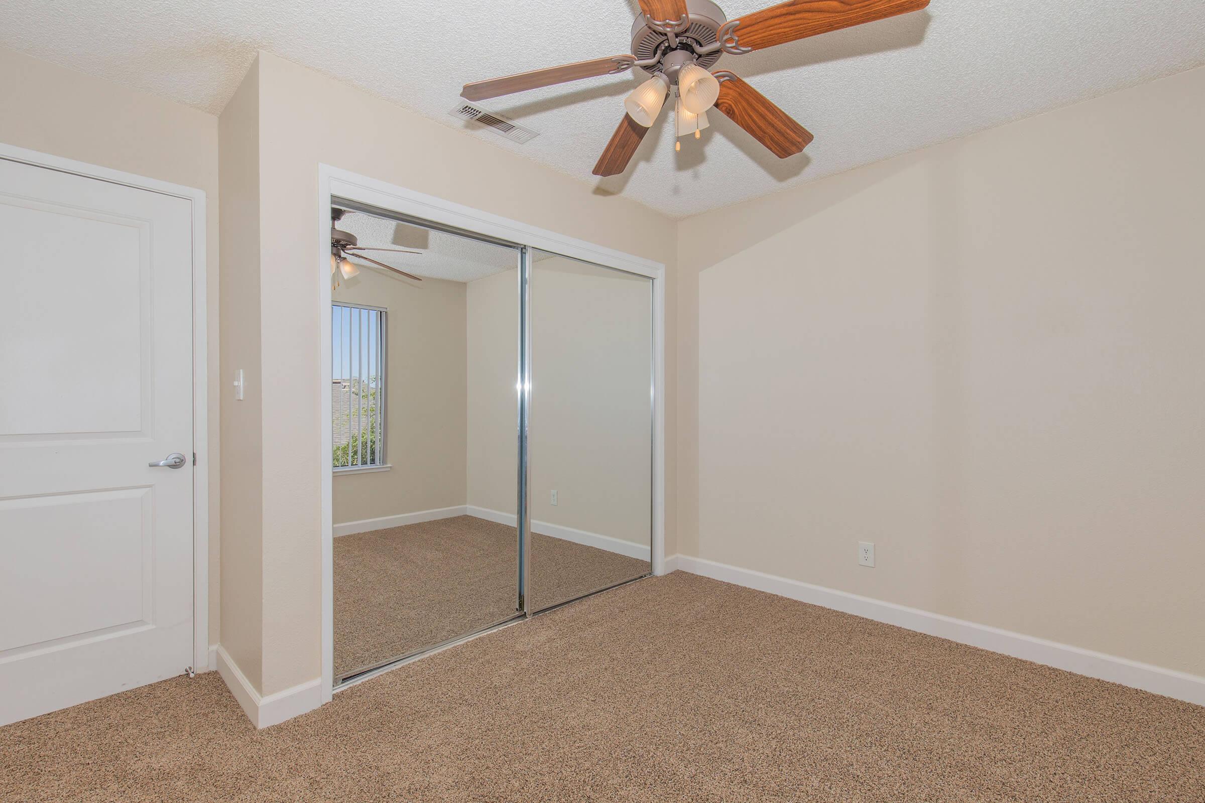 A well-lit room featuring beige walls and carpet flooring, with a ceiling fan having wooden blades. There are mirrored sliding closet doors on one wall and a window with blinds, allowing natural light to enter the space. The overall ambiance is neutral and inviting.