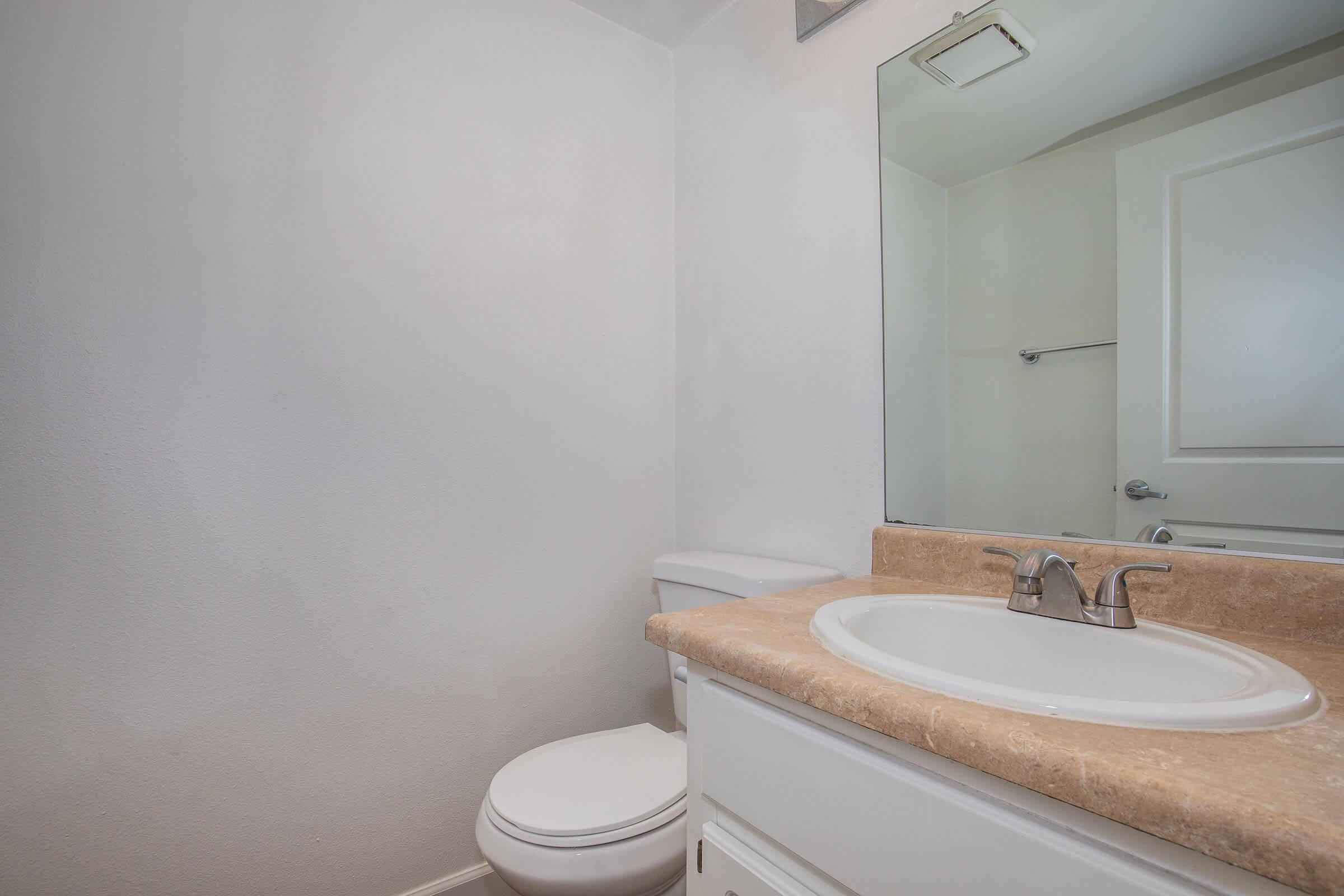 A clean, modern bathroom featuring a white toilet, a beige countertop sink with a chrome faucet, a large mirror above the sink, and plain white walls. The space is well-lit and minimalistic, with a towel rack visible in the background.