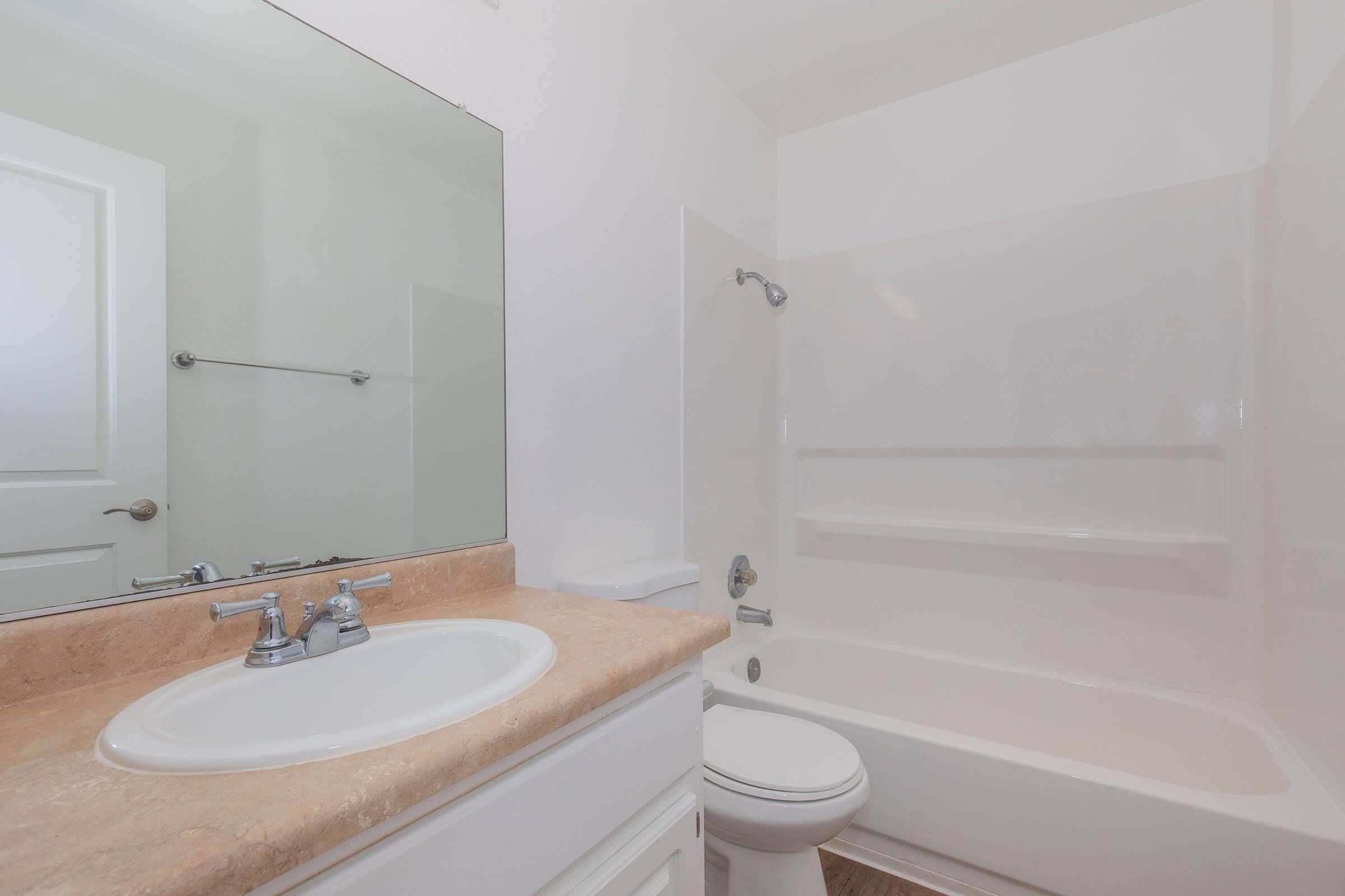 A clean, modern bathroom featuring a bathtub with a shower, a white toilet, and a sink with a countertop. A large mirror is mounted above the sink, and there are white walls, creating a bright and airy atmosphere. Simple and minimalist design with a focus on functionality.