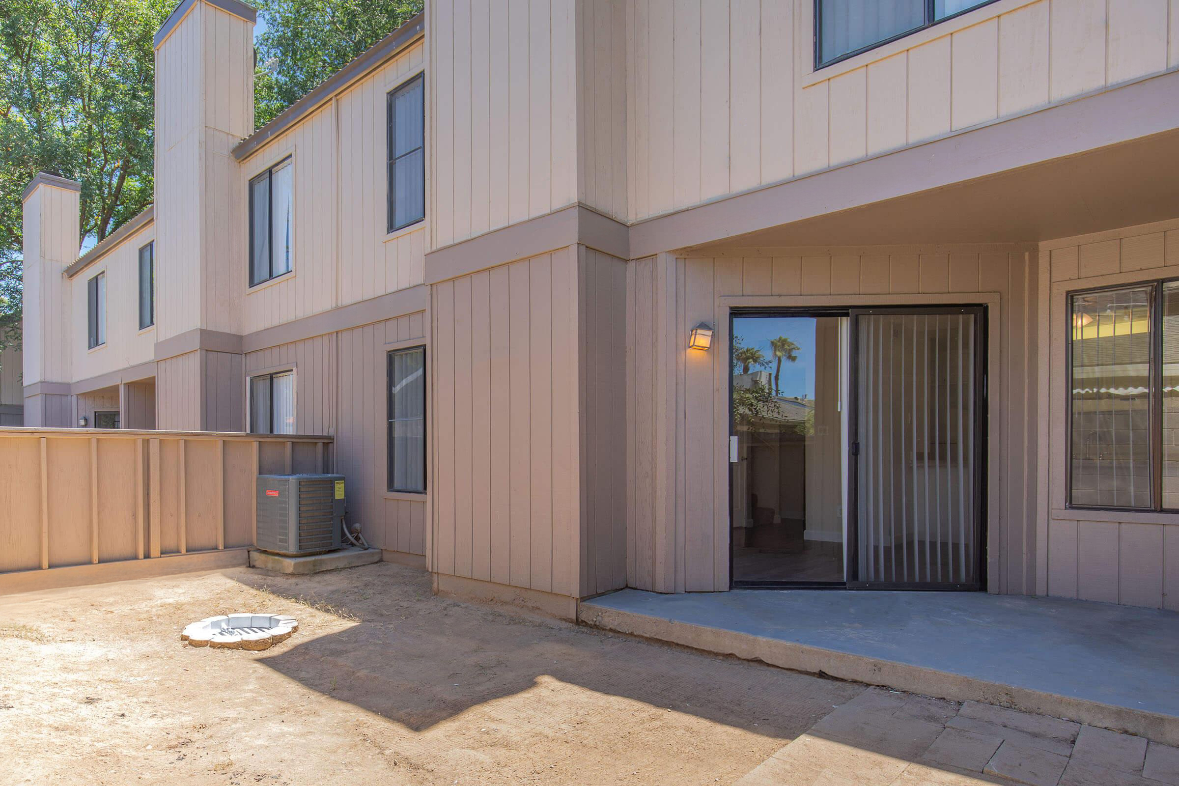 Exterior view of a modern residential building featuring beige siding, a sliding glass door with a vertical blind, and a small outdoor area with dirt and patio stones. An air conditioning unit is situated beside the entrance, and trees are visible in the background.