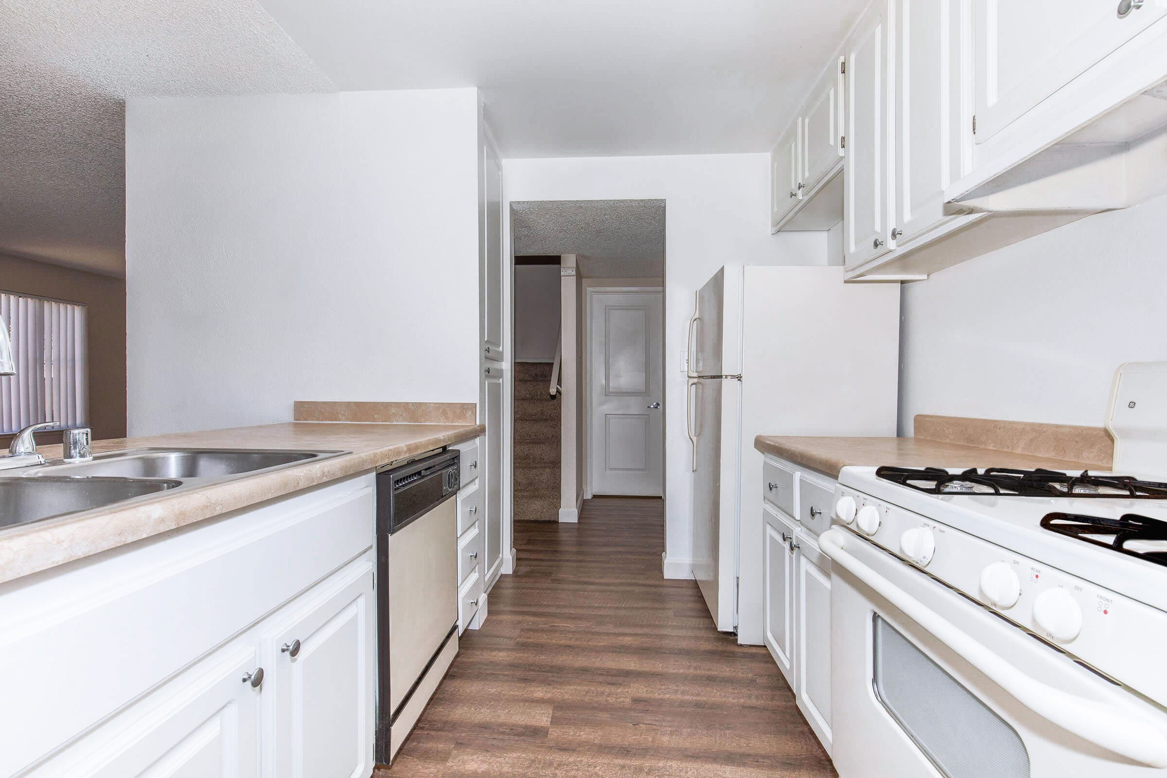 A modern kitchen featuring white cabinets, a double sink, and a gas stove. The countertop is light-colored, and there’s a dishwasher next to the sink. The space has vinyl flooring and a doorway leading to a staircase, with natural light coming in from a window.
