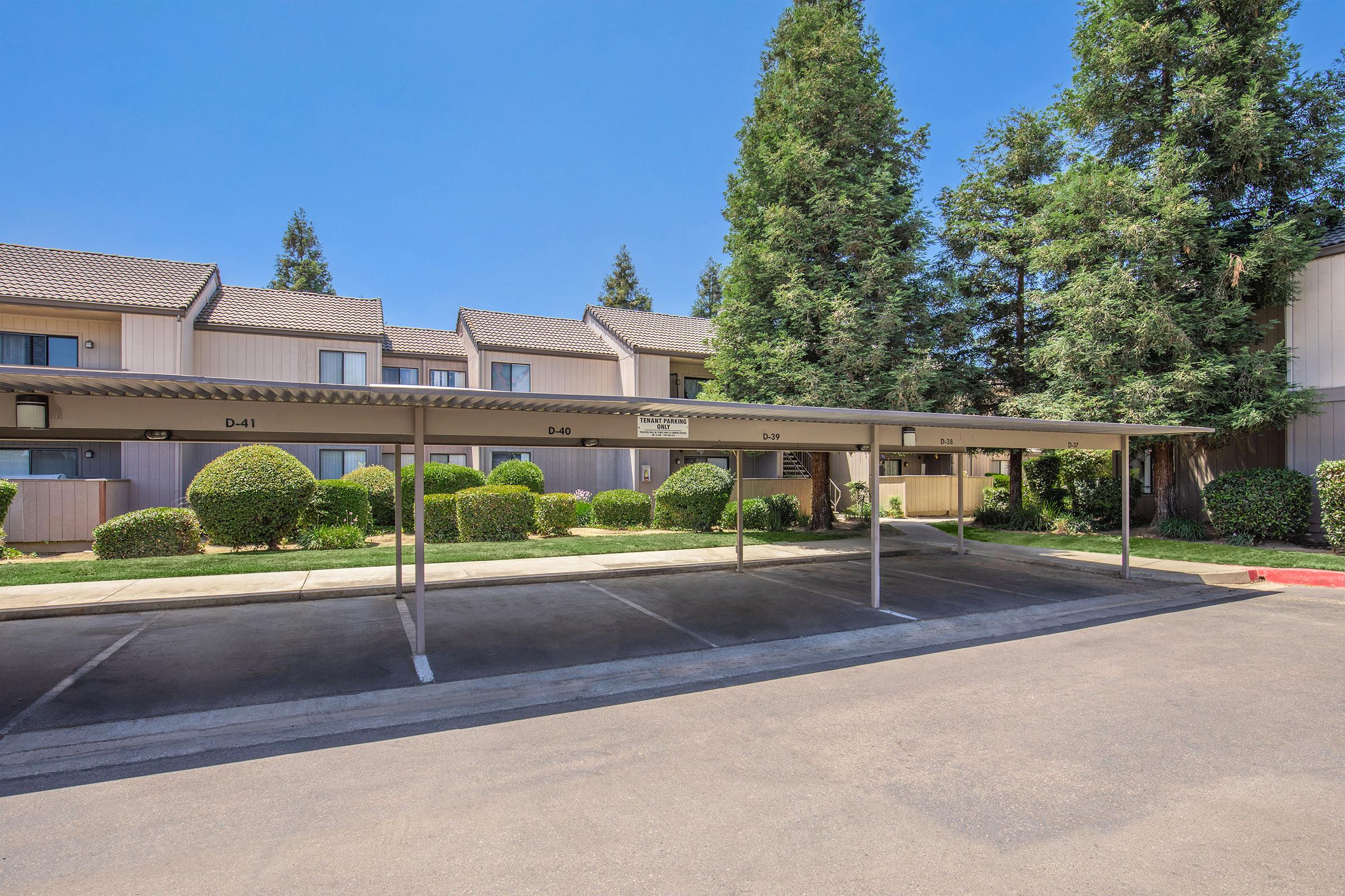 A parking area with covered spaces in front of an apartment complex. The scene features well-maintained green shrubs and trees, with a clear blue sky above. The buildings in the background are two stories tall, with balconies and a tidy appearance.
