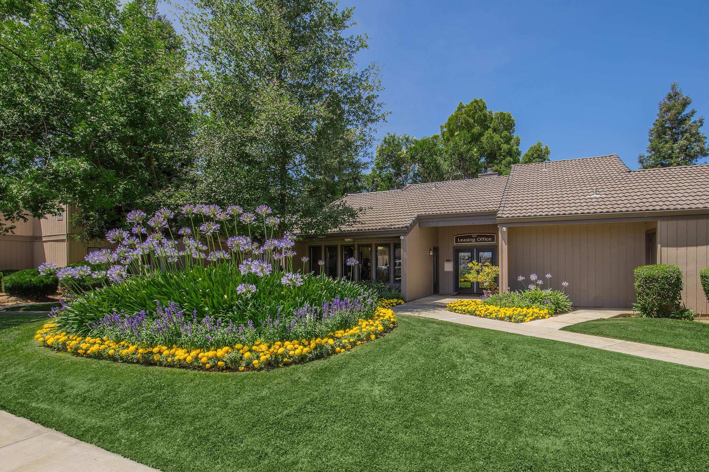 A well-maintained building entrance surrounded by vibrant flower beds featuring yellow and purple blooms. Lush green grass and trees provide a natural backdrop under a clear blue sky. The pathway leads to the entrance, creating an inviting and picturesque scene.