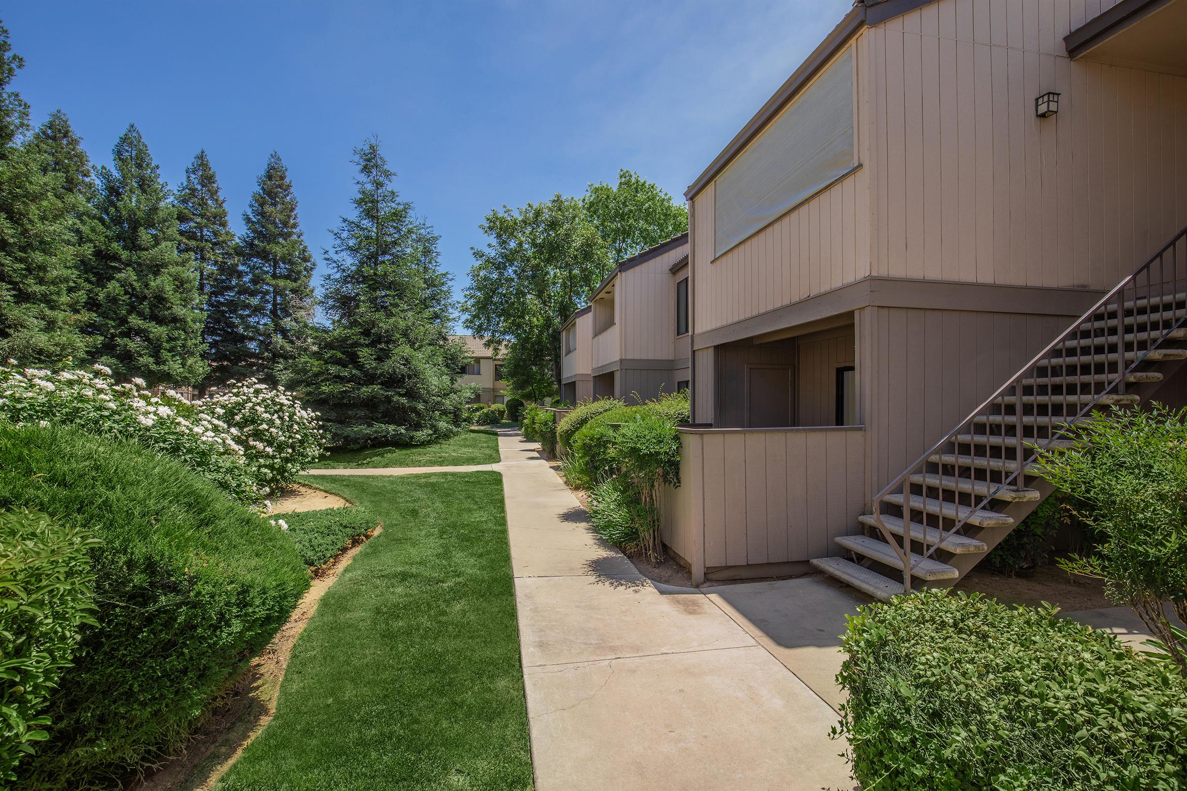 A landscaped pathway leading through a residential apartment complex. The path is flanked by well-maintained greenery, including shrubs and trees. A staircase leads to the upper level of an apartment building on the right, under a clear blue sky. The scene evokes a serene and inviting atmosphere.