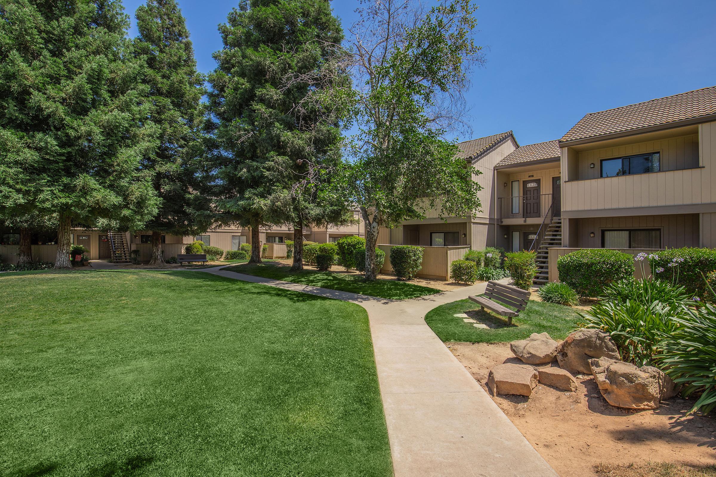 A landscaped courtyard with green grass, trees, and shrubs. Pathways lead between multi-story residential buildings. A bench is visible on the left, and there are rocks and flower beds in the foreground, set against a clear blue sky.