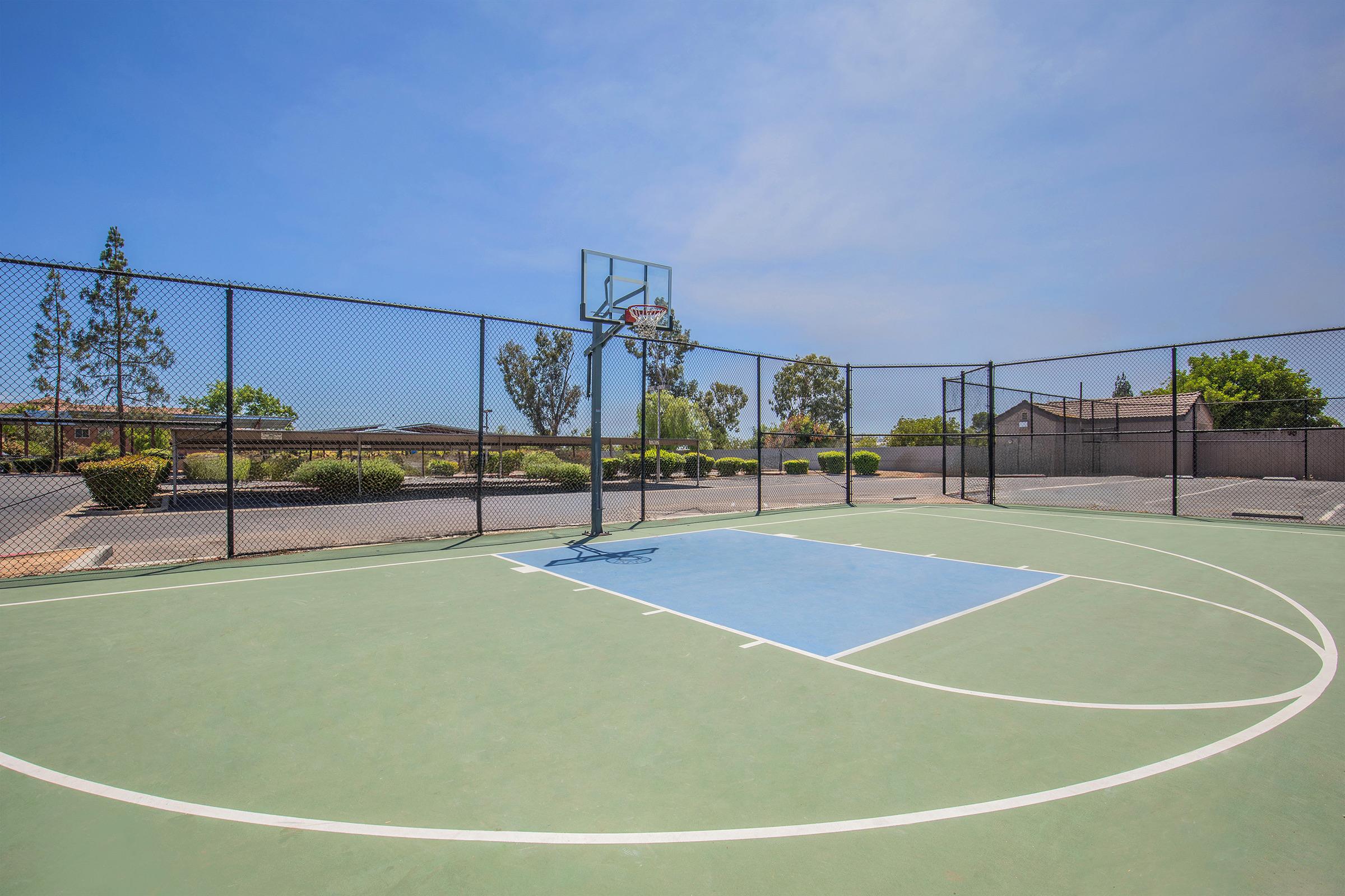 A view of an outdoor basketball court featuring a green surface, a blue-painted section near the hoop, and a chain-link fence surrounding the area. In the background, there are trees and parked cars, under a clear blue sky. The scene conveys a peaceful, sunny day ideal for playing basketball.