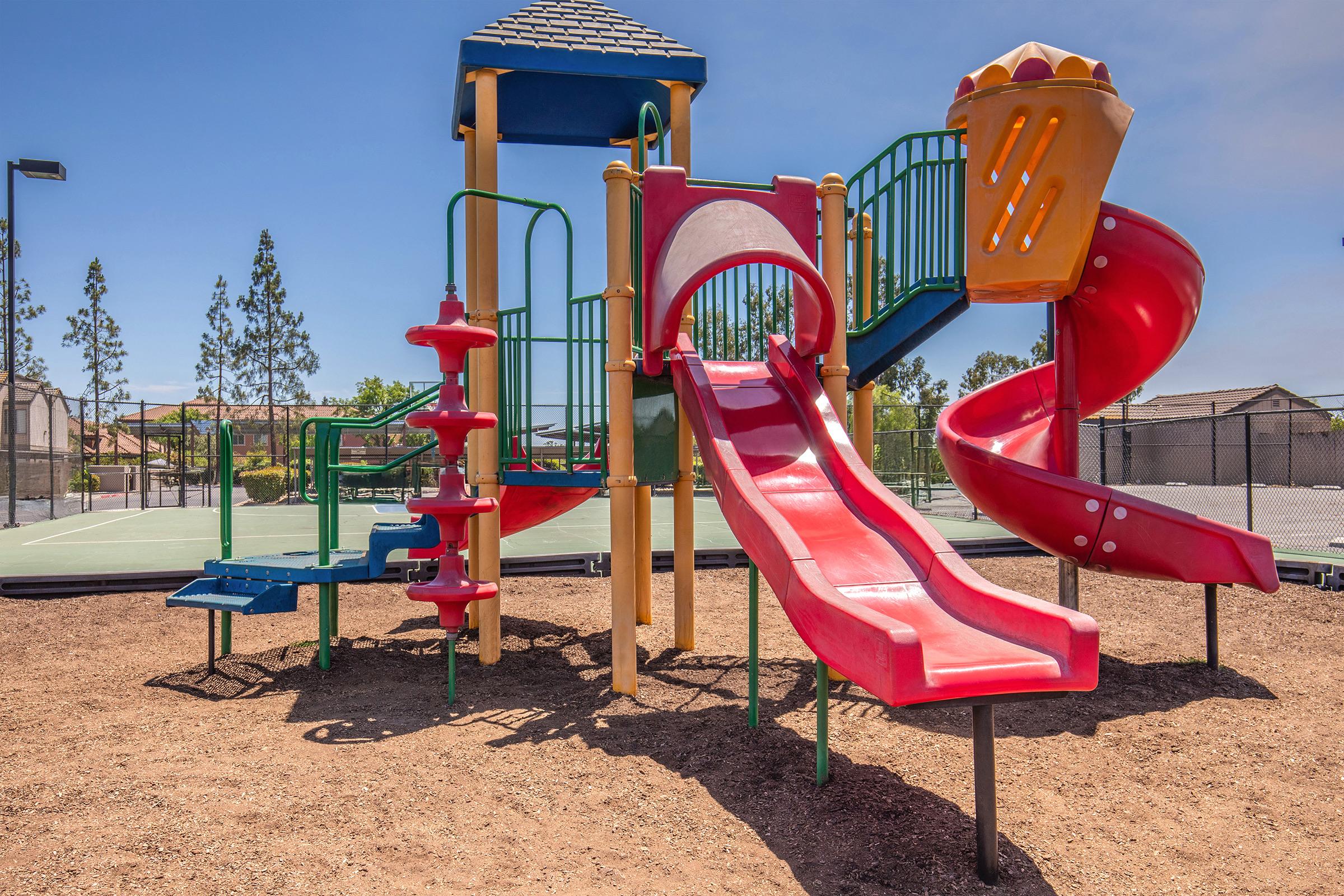 Colorful playground structure featuring two slides, one straight and one curved, alongside climbing features. The playground is surrounded by a sandy area and has a basketball court in the background under a clear blue sky.
