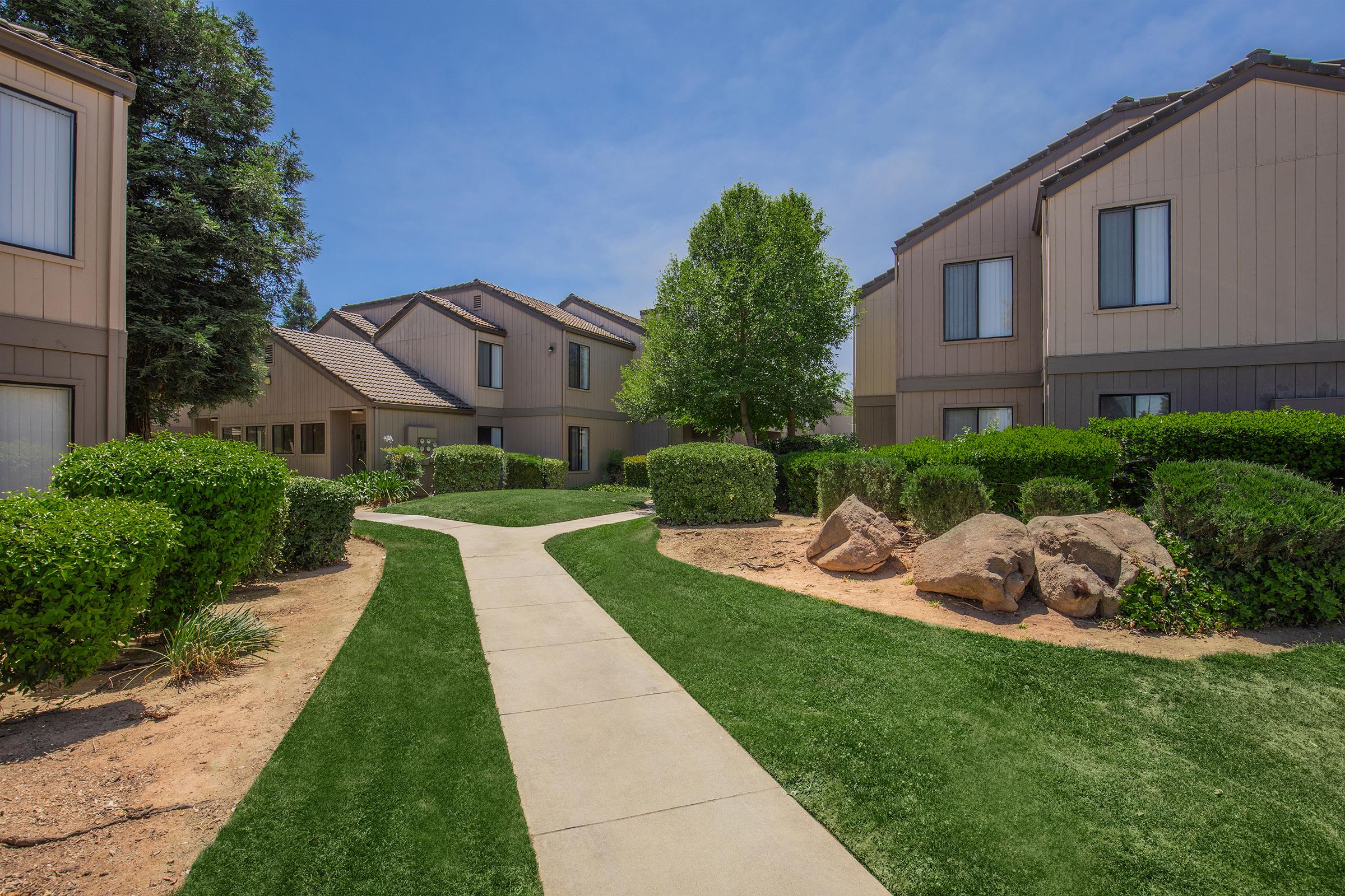 A well-maintained pathway winds through a landscaped area between two residential buildings. Lush green shrubs and bushes line the path, with a few large rocks and patches of grass enhancing the natural beauty of the scene. Bright blue sky adds to the inviting atmosphere.
