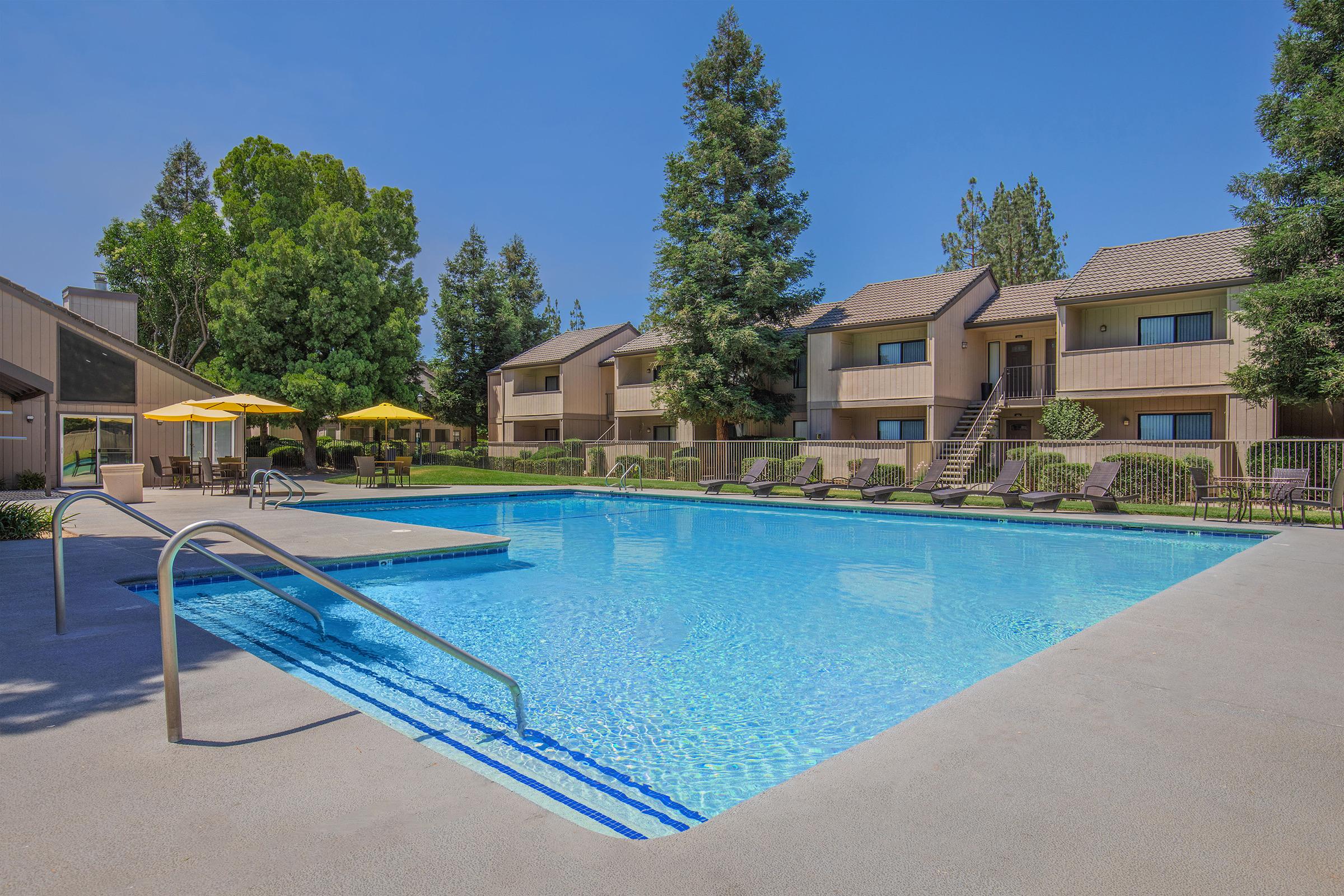 A clear blue swimming pool surrounded by lounge chairs and shaded by yellow umbrellas. In the background, there are trees and two-story apartment buildings with balconies. The scene reflects a sunny day, creating a relaxing outdoor atmosphere.