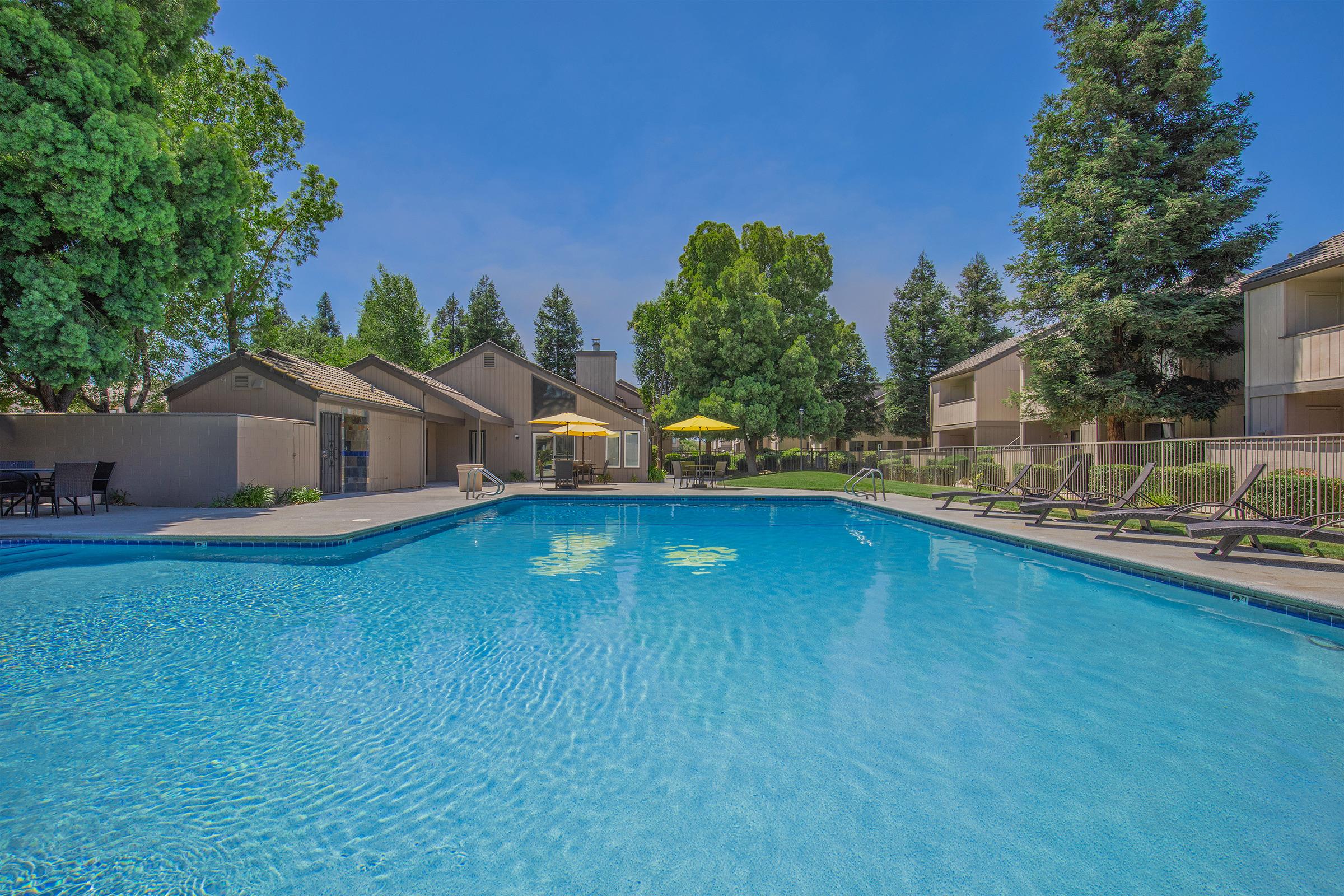 Swimming pool surrounded by lounge chairs, with a few yellow umbrellas providing shade. Lush green trees and neatly landscaped grounds are visible in the background, along with residential buildings. Clear blue skies create a peaceful and inviting atmosphere.