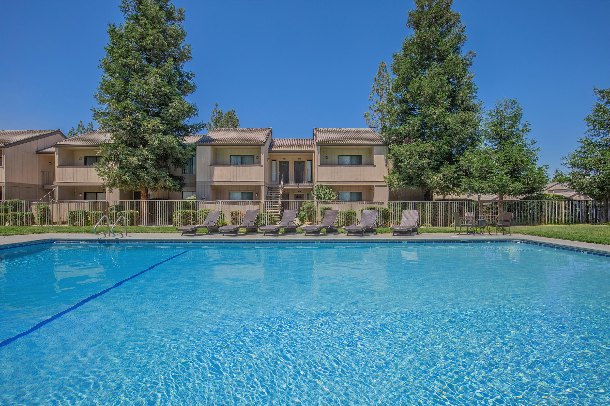 A clear blue swimming pool surrounded by lounge chairs, with green grass and tall trees in the background. There are two-story apartment buildings visible behind the pool, under a bright blue sky.