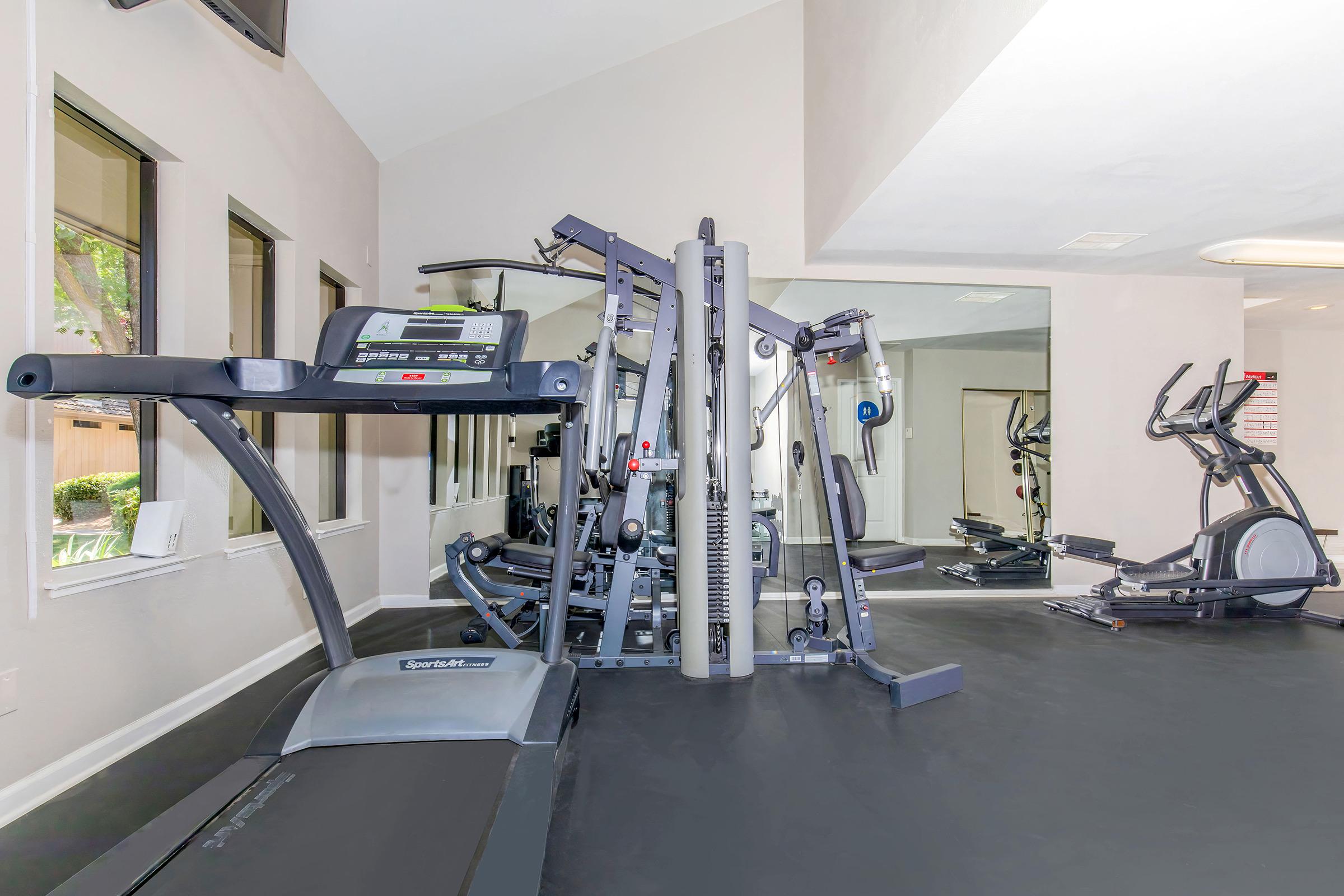 Modern gym interior featuring a treadmill, multi-station weight machine, and an elliptical trainer. The space is well-lit with large windows, creating an inviting atmosphere for workouts. The floor is black, enhancing the sleek design of the fitness equipment.