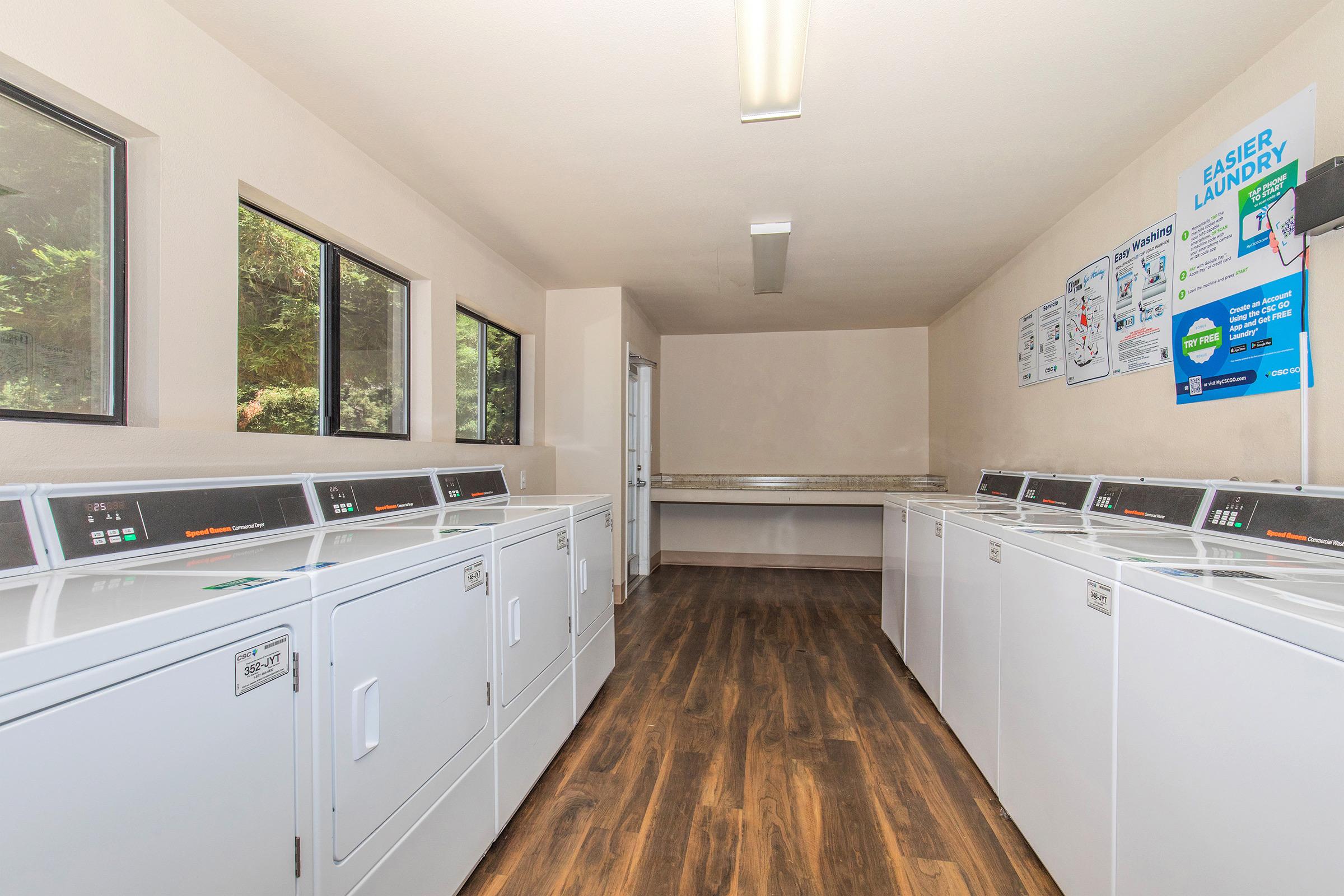 A clean, well-lit laundry room featuring several rows of white washing machines and dryers. Large windows let in natural light, and there is a counter space at one end. Posters on the wall provide laundry tips and instructions. The flooring is a warm wood finish.