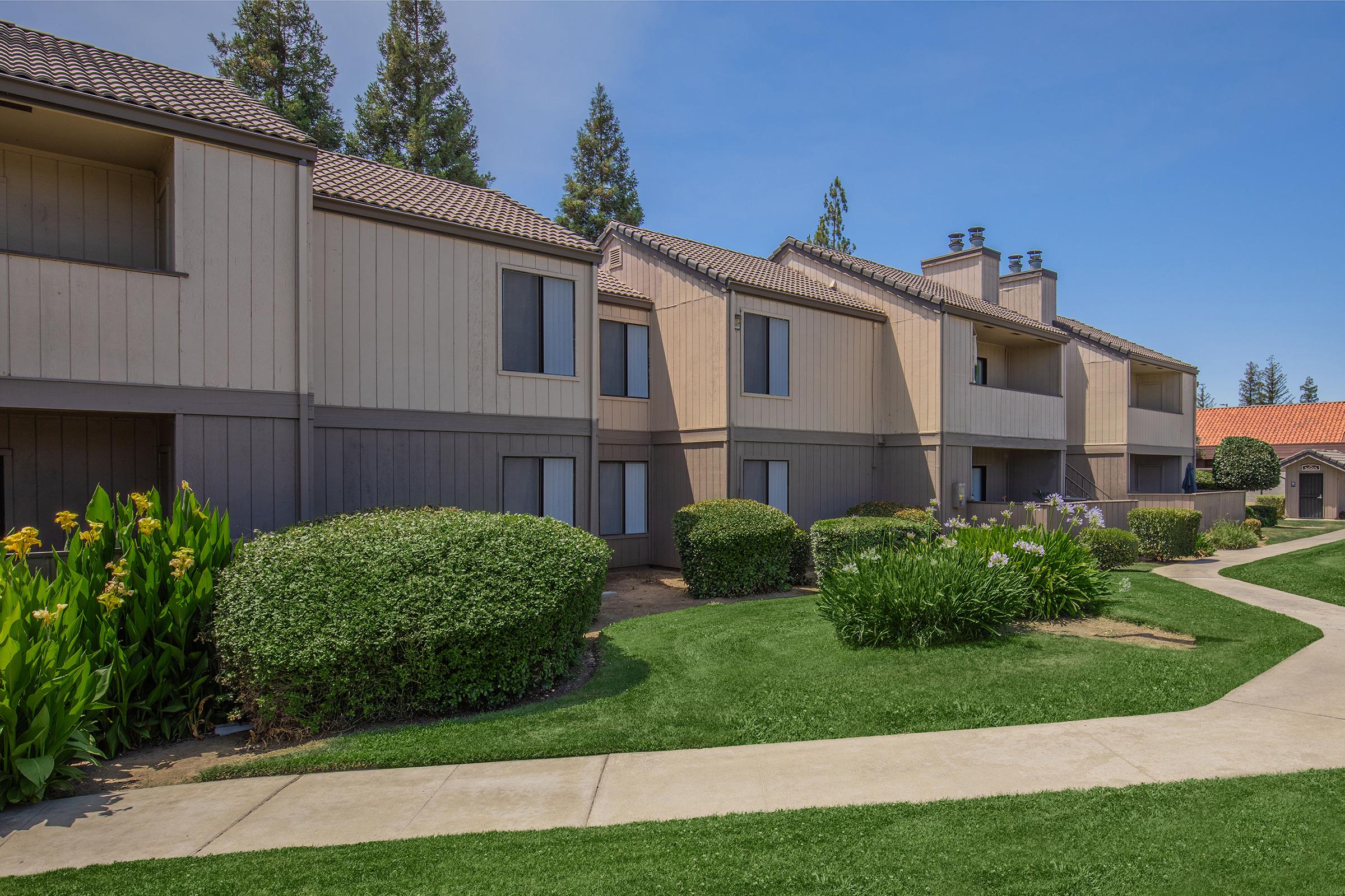 A view of two-story apartment buildings with a light brown exterior, surrounded by well-maintained green lawns and shrubs. Pathways wind through the landscape, leading towards the buildings under a clear blue sky. Trees are visible in the background.
