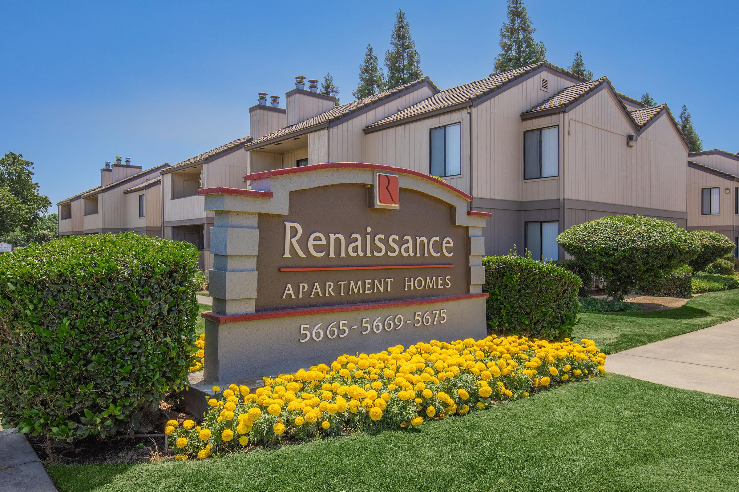 Sign for Renaissance Apartment Homes with colorful flower beds in front. The building features a light exterior with multiple units visible in the background, surrounded by neatly trimmed bushes and trees under a clear blue sky.