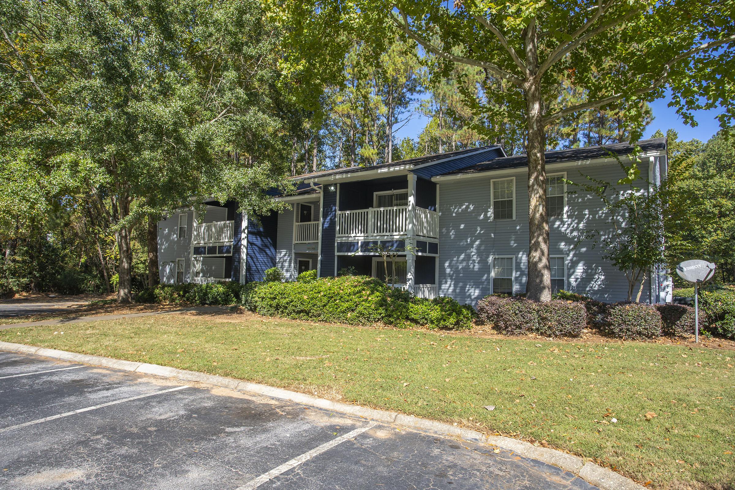 A two-story apartment building surrounded by green trees and a well-maintained lawn. The building features balconies on the upper level and is partially shaded by large trees. A parking lot with asphalt is visible in the foreground, with a few parked cars and a satellite dish in the yard.