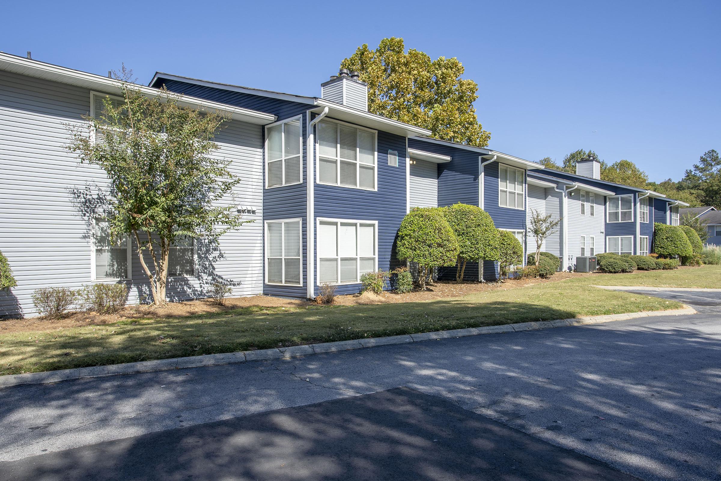 Exterior view of a multi-unit apartment building featuring a mix of light blue and dark blue siding. The landscape includes neatly trimmed bushes and trees, with a clear blue sky in the background. The scene is well-maintained, showing a paved pathway and a green lawn.