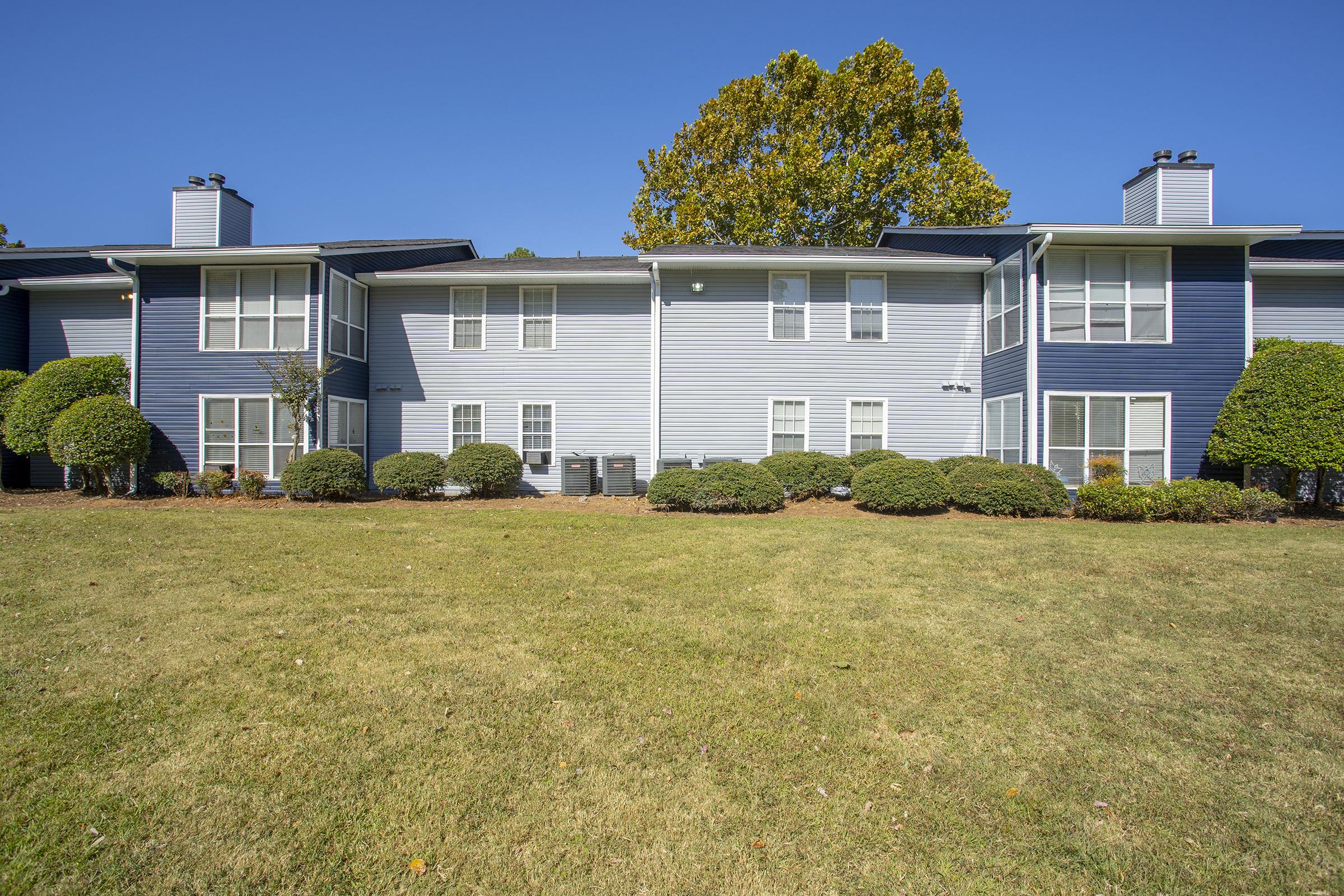 Two-story apartment building with blue siding and multiple windows. The landscaping includes neatly trimmed bushes in front and a large grassy area. The sky is clear and blue, and there are trees in the background.