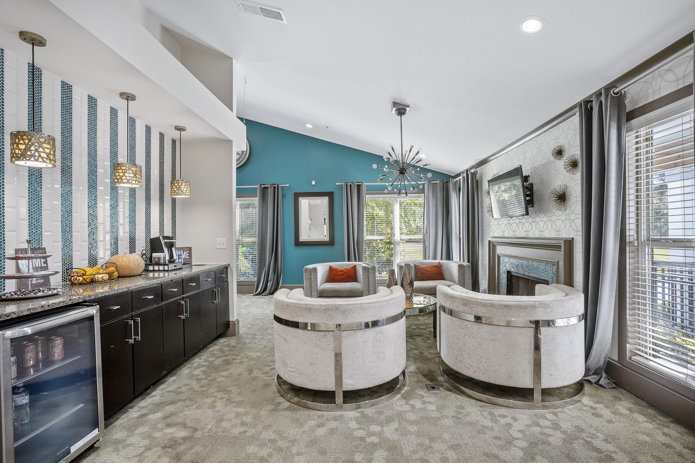 A modern living space featuring two stylish chairs with light-colored upholstery, a chic coffee table, and a dark wood cabinet with a mini bar. The walls are decorated with a blue accent wall and patterned wallpaper, while large windows allow natural light. There’s also a decorative chandelier and contemporary lighting fixtures.