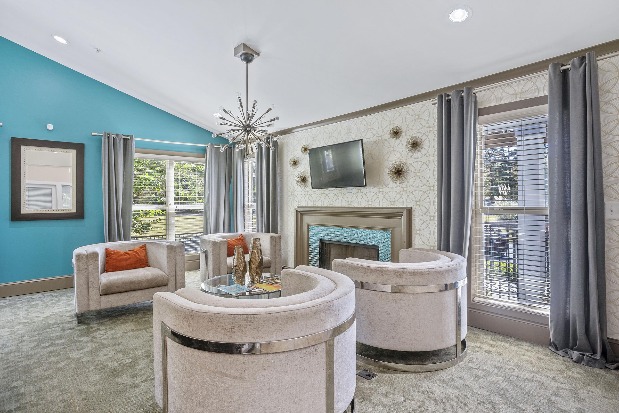 A modern living room featuring two stylish white armchairs and a cozy fireplace with a teal accent wall. Large windows with gray curtains allow natural light in, while a minimalist coffee table holds decorative items. A wall-mounted TV is positioned above the fireplace, adding to the contemporary feel.