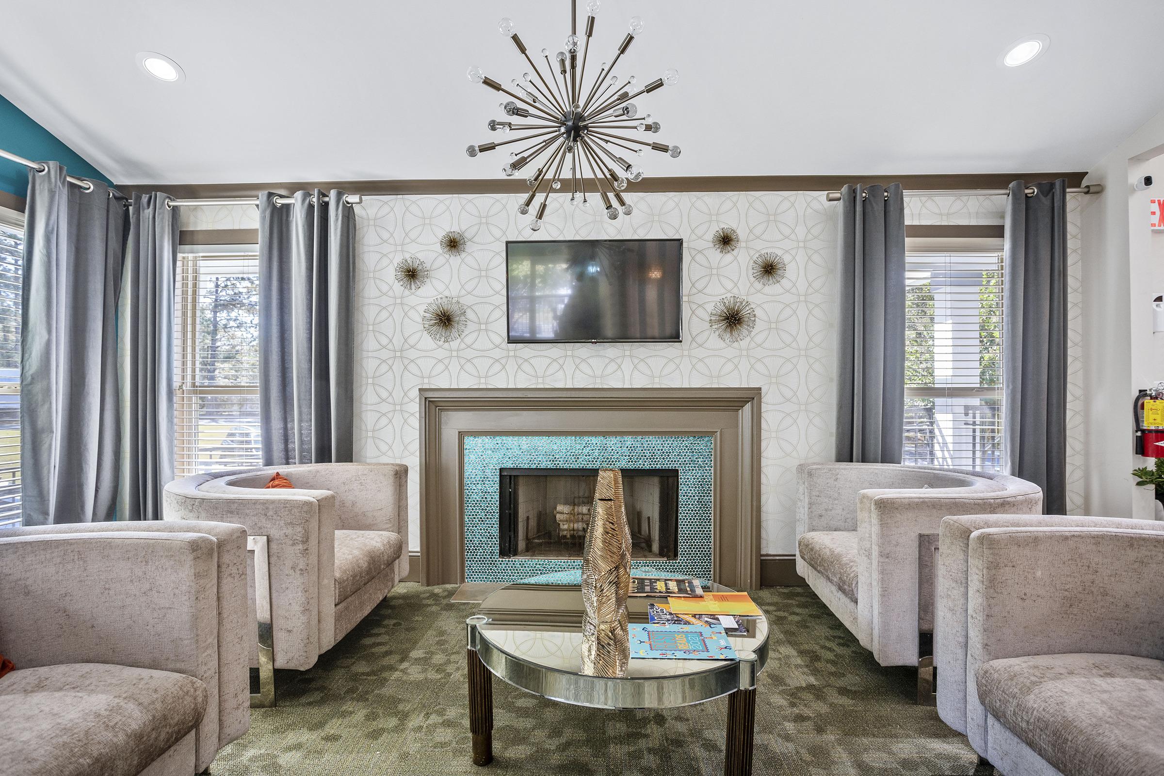 Cozy lounge area featuring modern furniture with four plush armchairs, a decorative coffee table with books, and a stylish gas fireplace. A large flat-screen TV hangs above the fireplace, and unique wall decor complements the bright, airy atmosphere created by ample natural light.